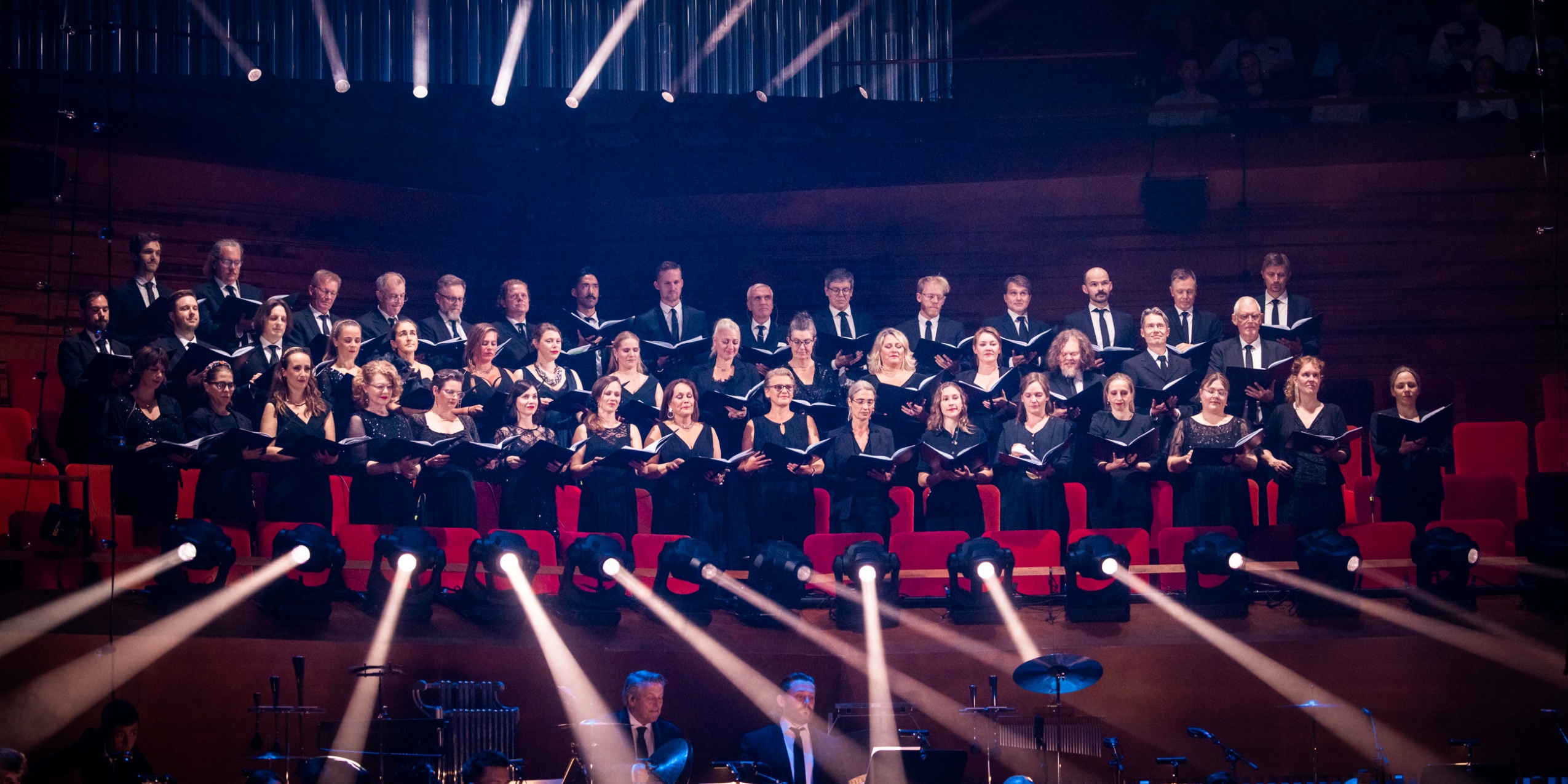Kor der synger i koncertsalen med orkesteret i forgrunden.