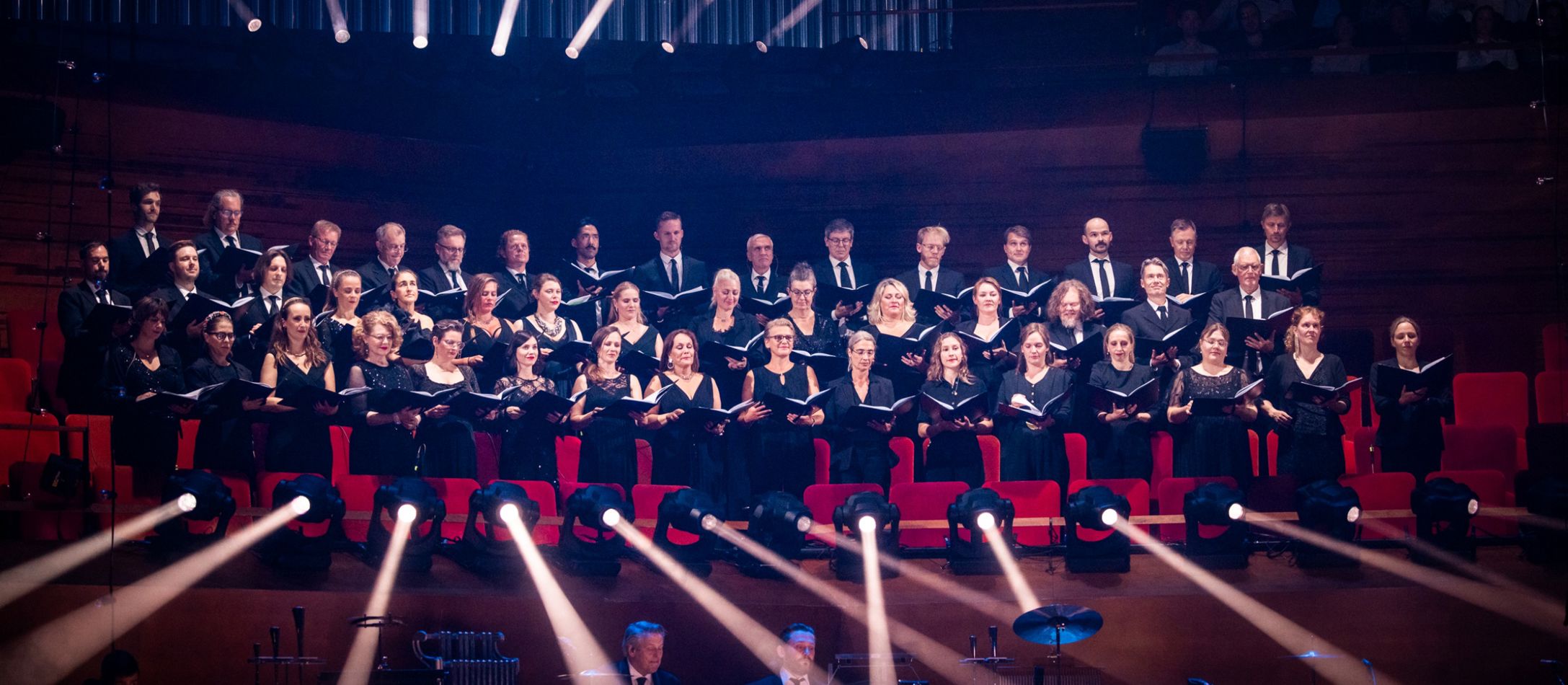 Kor der synger i koncertsalen med orkesteret i forgrunden.
