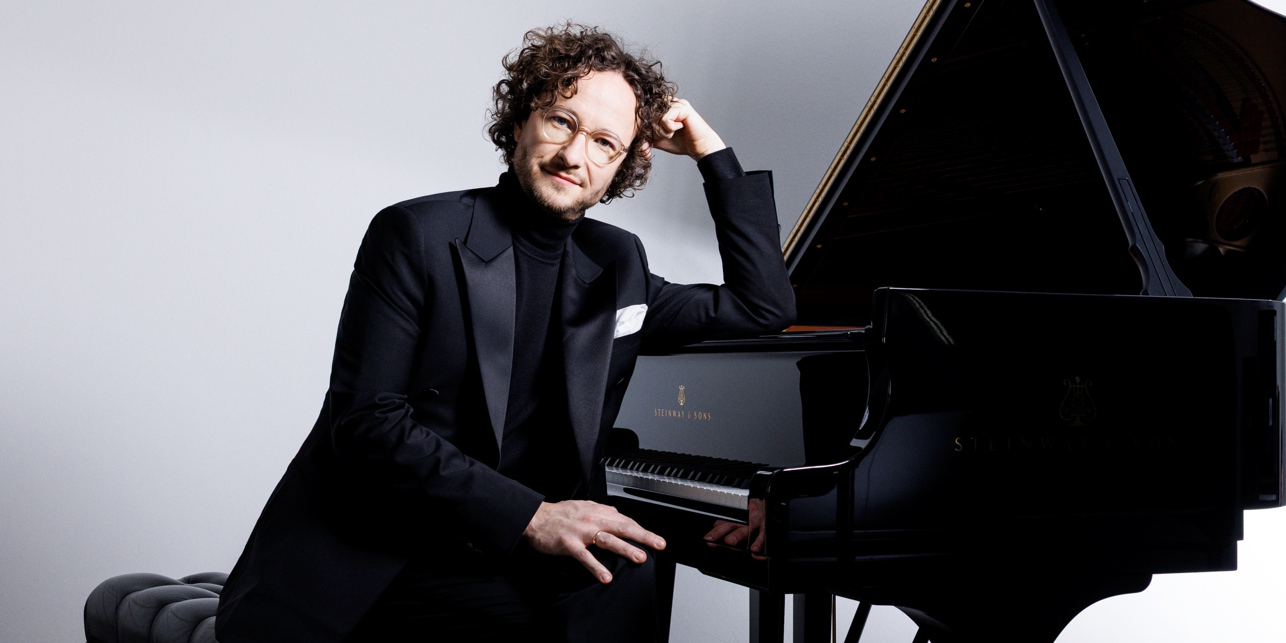 Mand i sort rullekrave og sort blazer siddende ved klaver med lys grå baggrund
