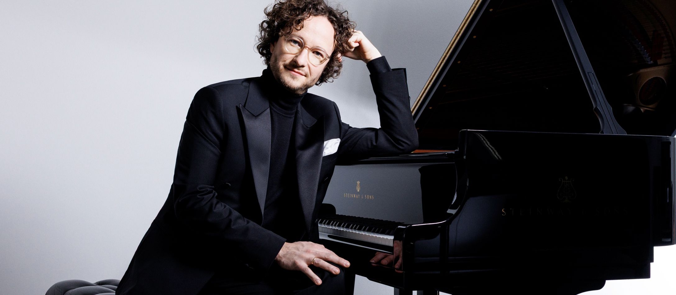 Mand i sort rullekrave og sort blazer siddende ved klaver med lys grå baggrund