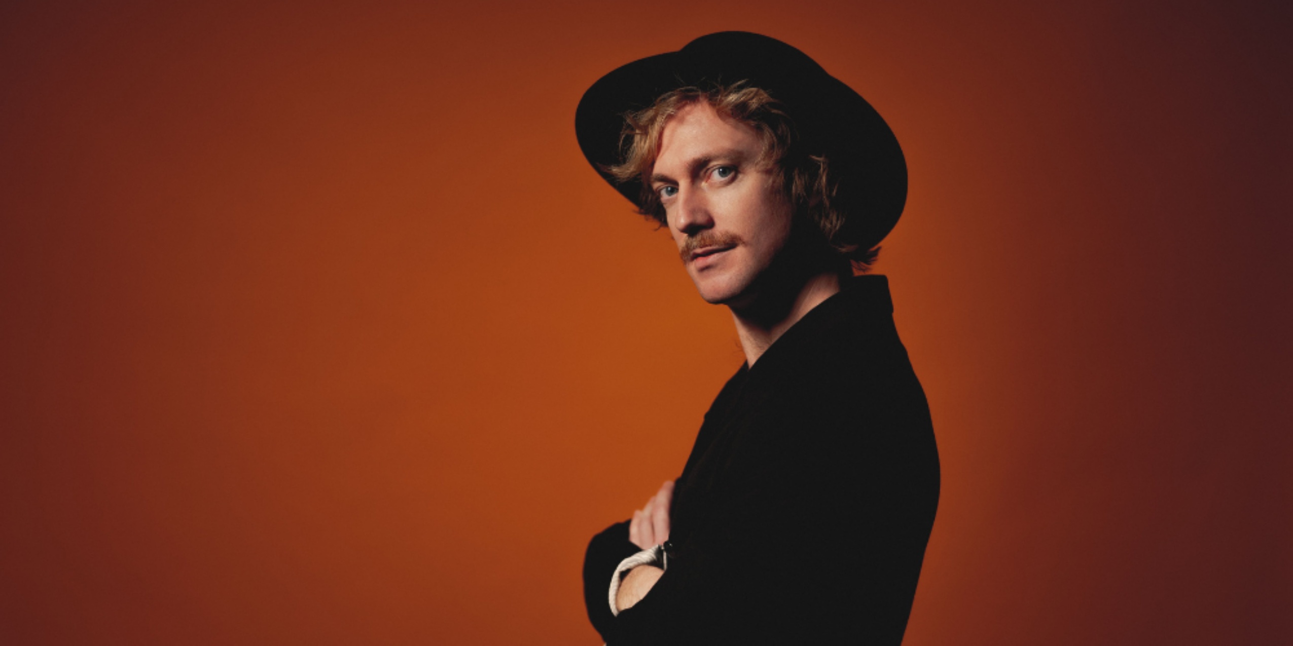 Mand i sort jakke og sort hat med stor skygge, står foran orange væg.