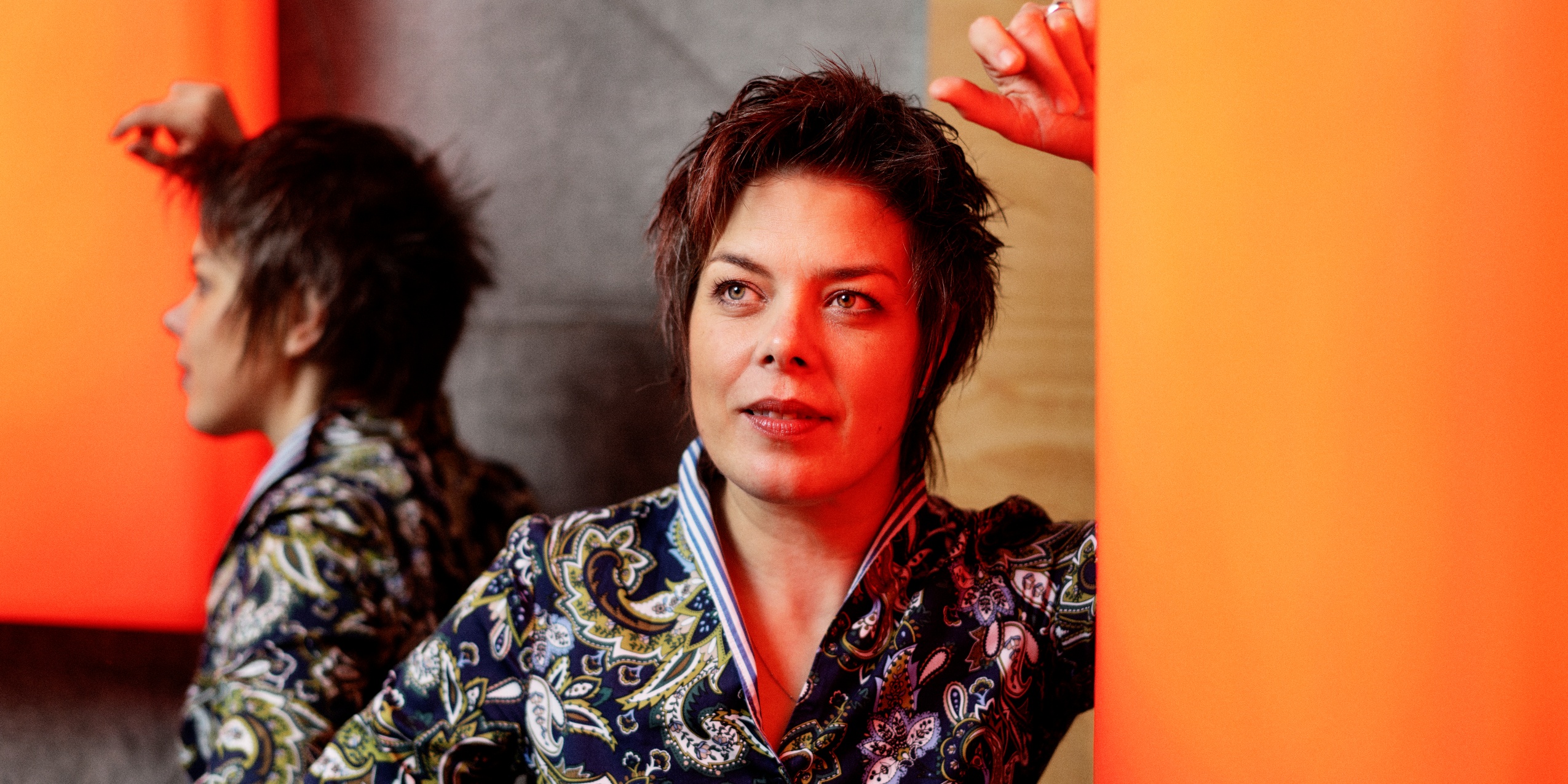 Kvinde i mønstret bluse, læner sig op ad en orange væglampe.