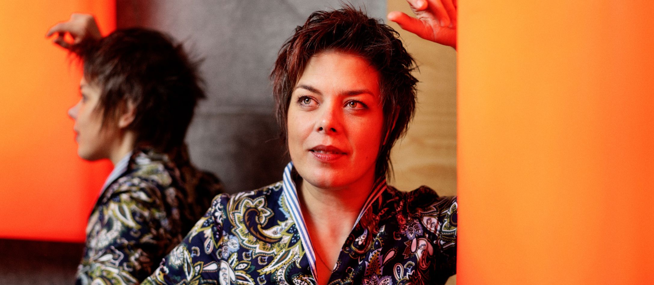 Kvinde i mønstret bluse, læner sig op ad en orange væglampe.