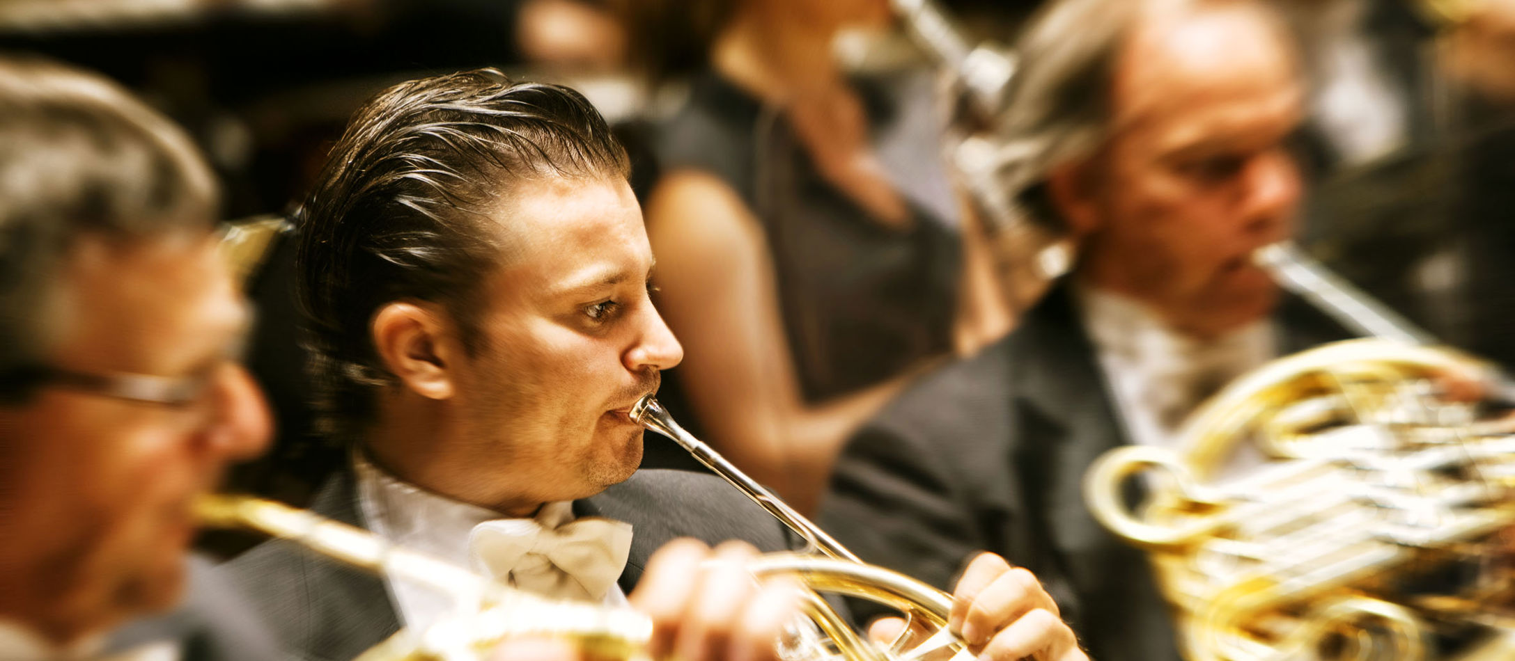 Lasse Mauritzen spiller horn til klassisk koncert iført klassisk jakkesæt og slikket hår ved siden af to kollegaer.