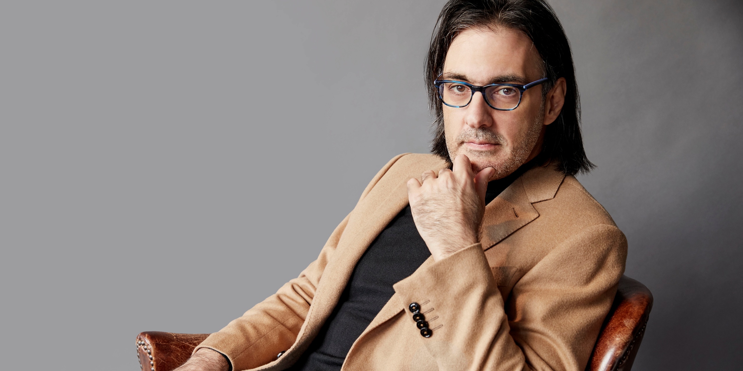 Mand i beige blazer, sidder i en stol foran grå væg.