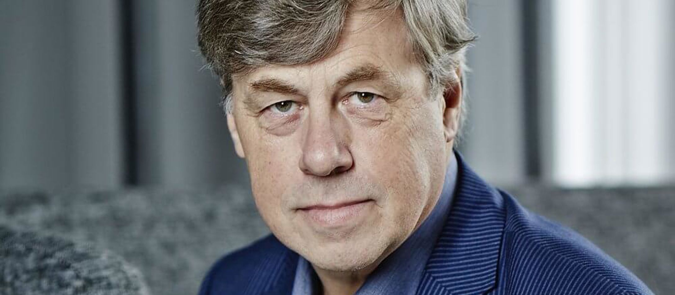 Siddende Paul Hillier med gråt hår kigger afslappet iført lyseblå skjorte og mørkeblå jakke.