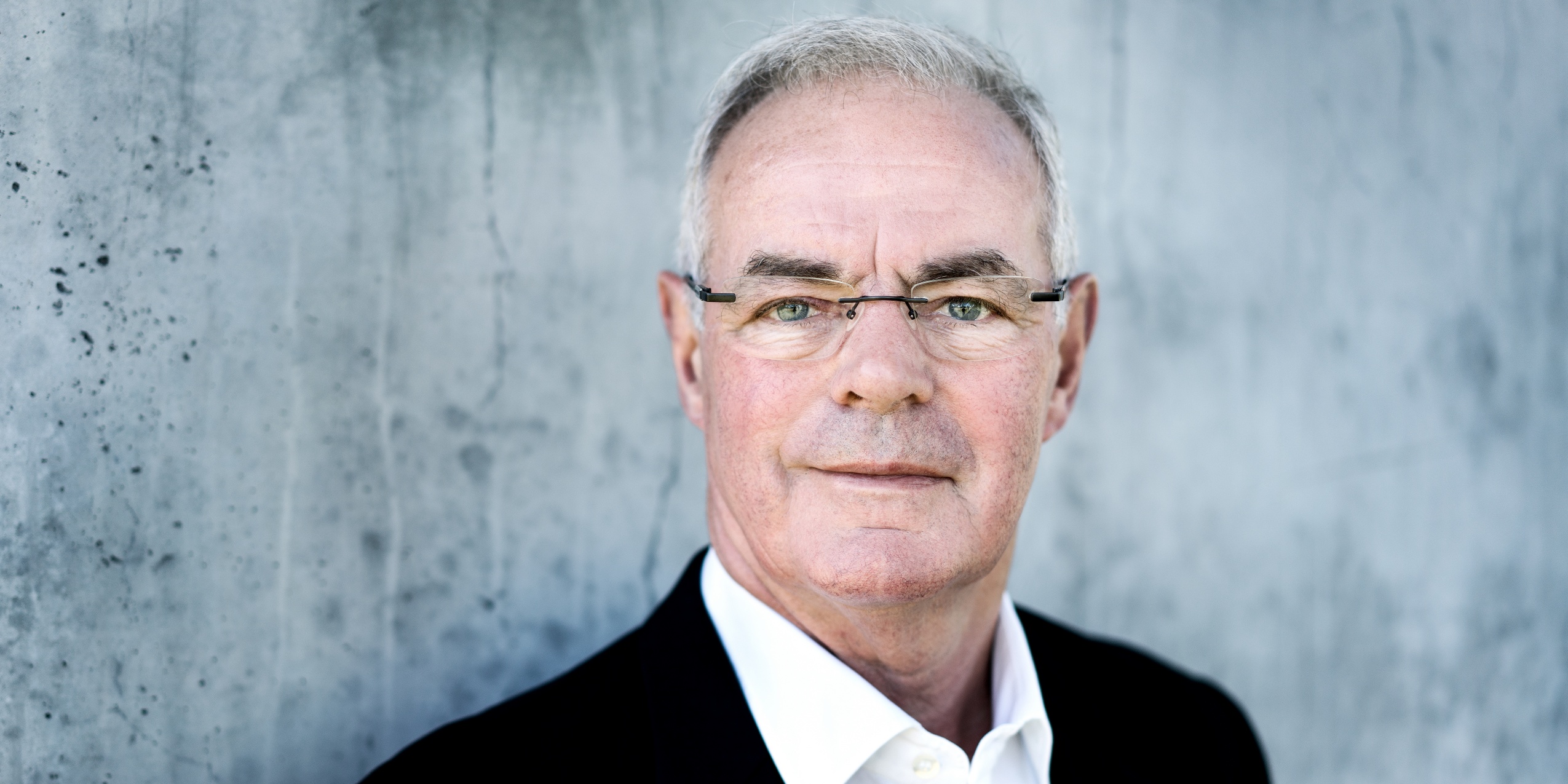 Mand i sort blazer og hvid skjorte, kigger ind i kameraet foran en betonvæg.