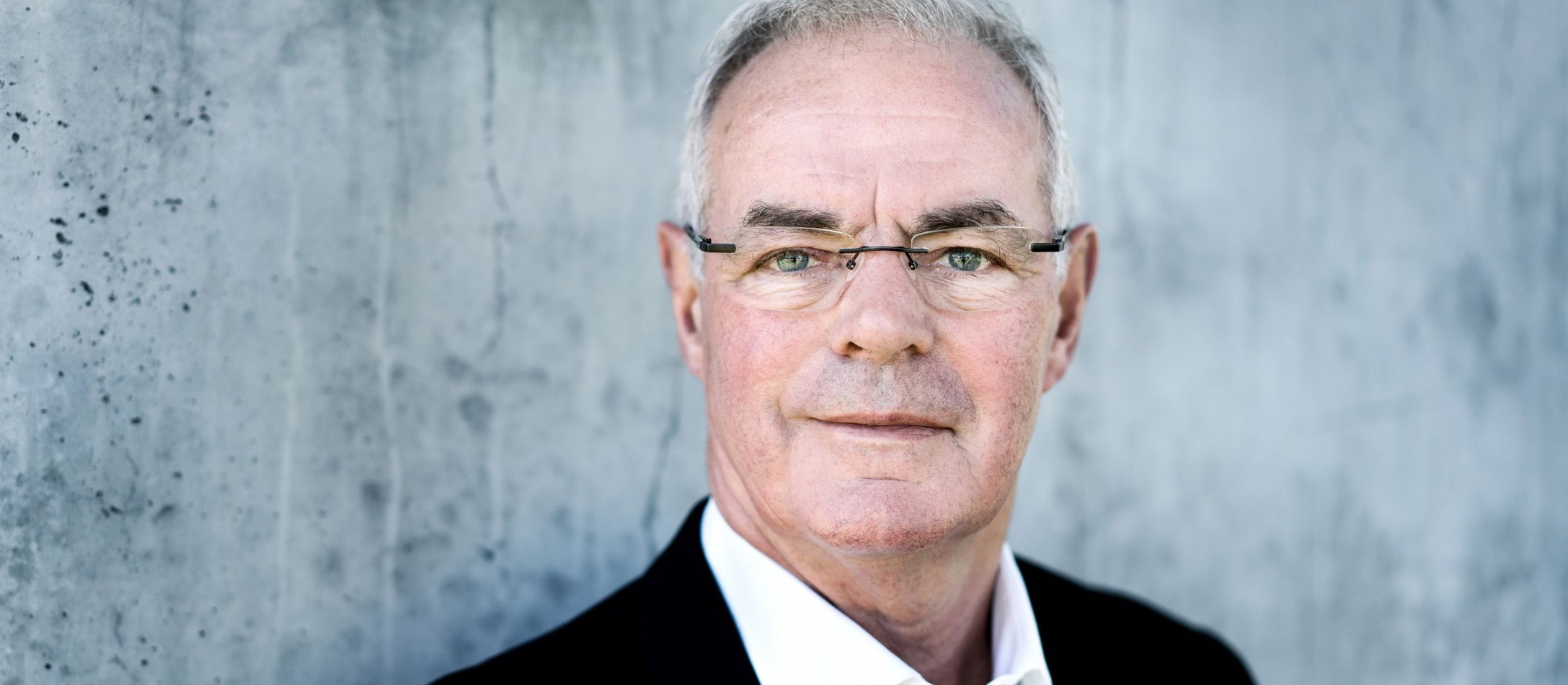 Mand i sort blazer og hvid skjorte, kigger ind i kameraet foran en betonvæg.