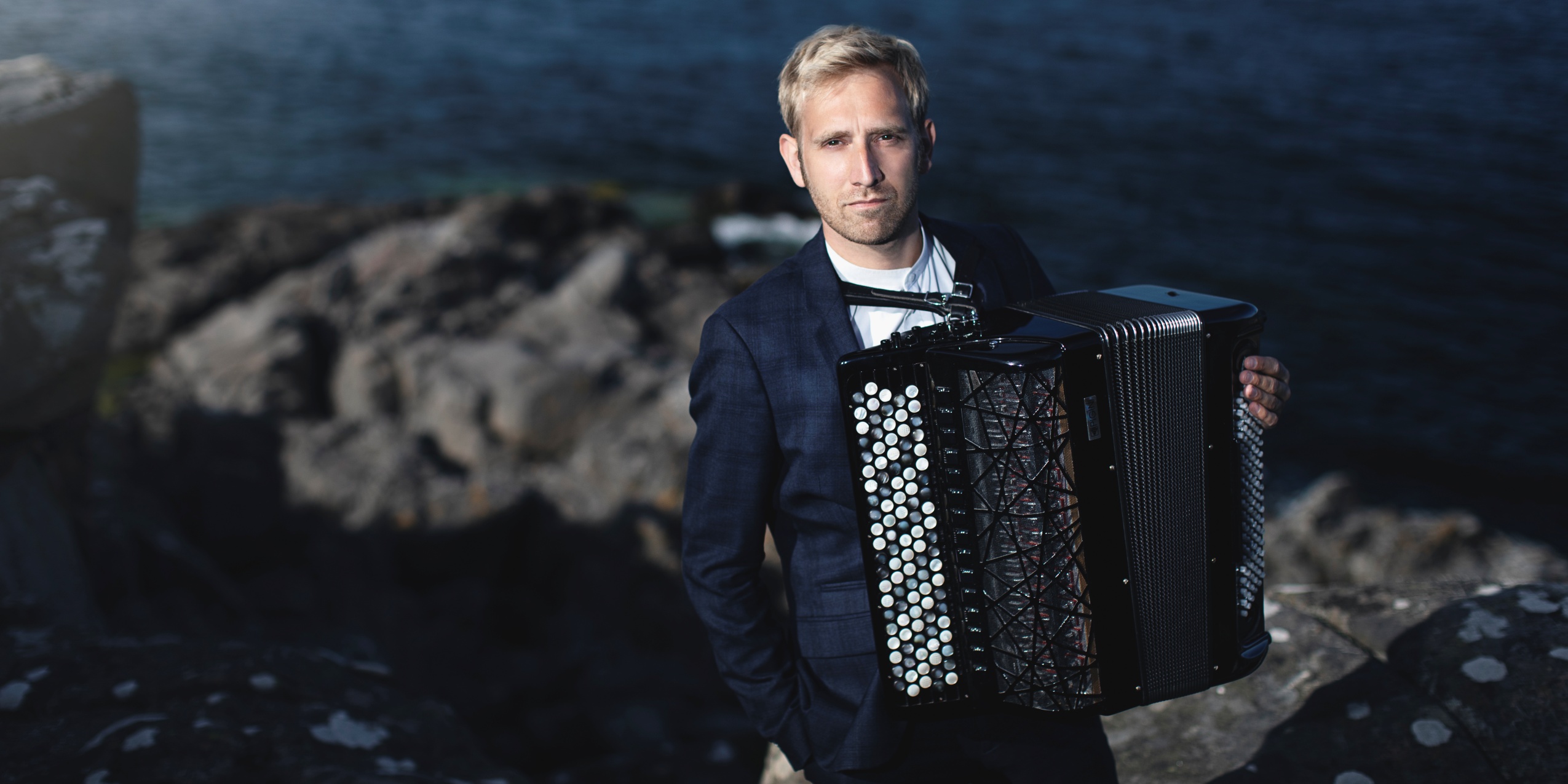 Mand med en akkordeon står på en klippeformation ved havet