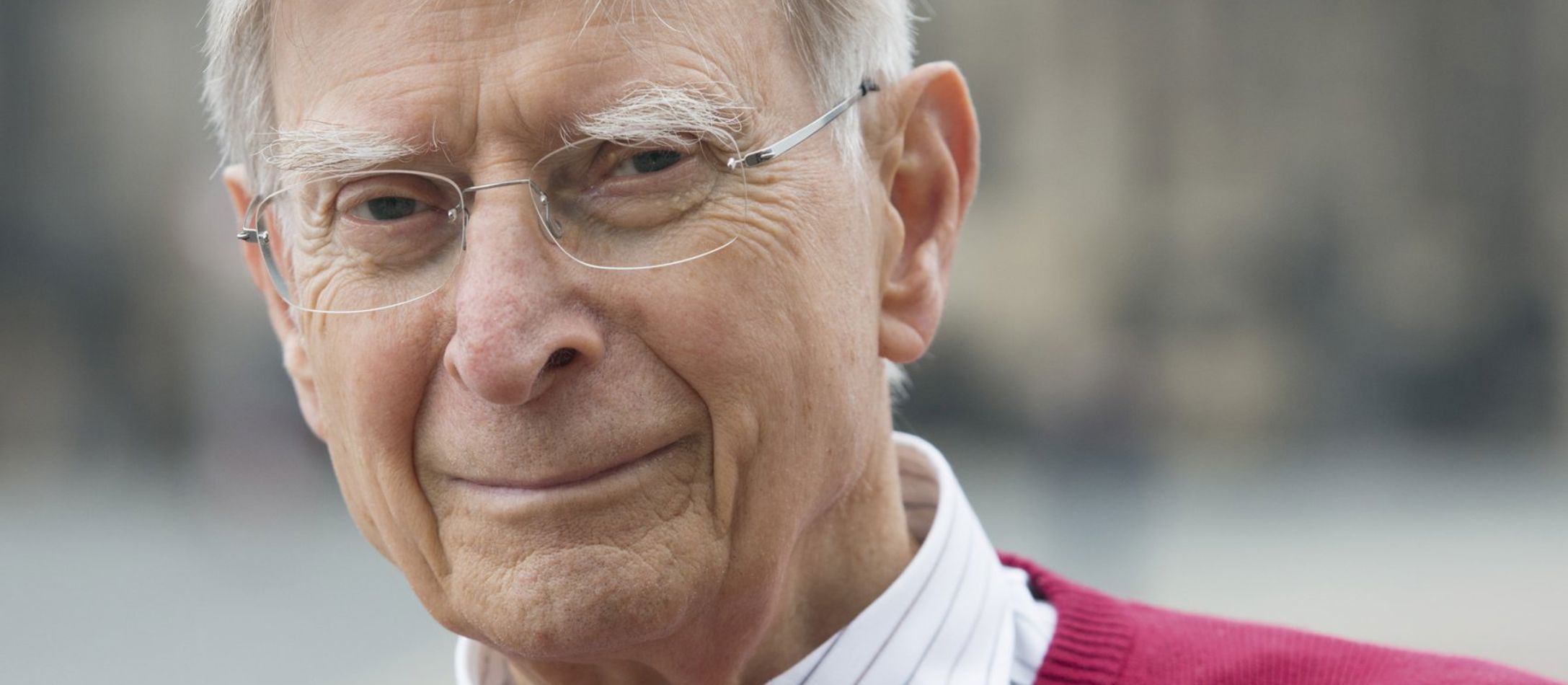 Herbert Blomstedt iført rød pullover med en hvidstribet skjorte med kortslagte arme foran sløret lys baggrund