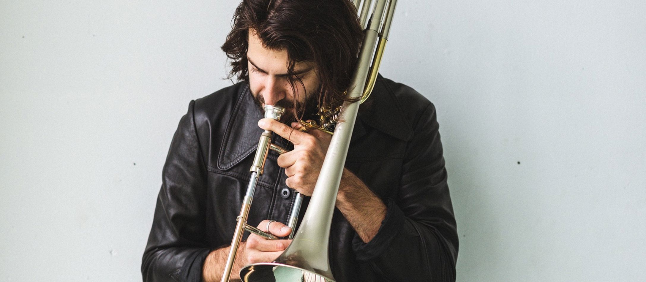 Mand der spiller basun i sort læderjakke foran hvid baggrund.