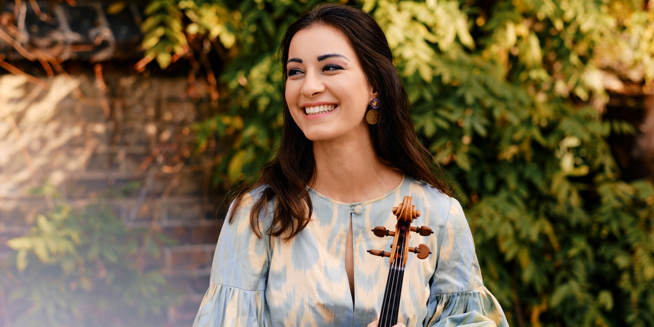 Kvinde i blå og hvid kjole, står smilende og holder sin bratsch, foran en mur og grønne grene.