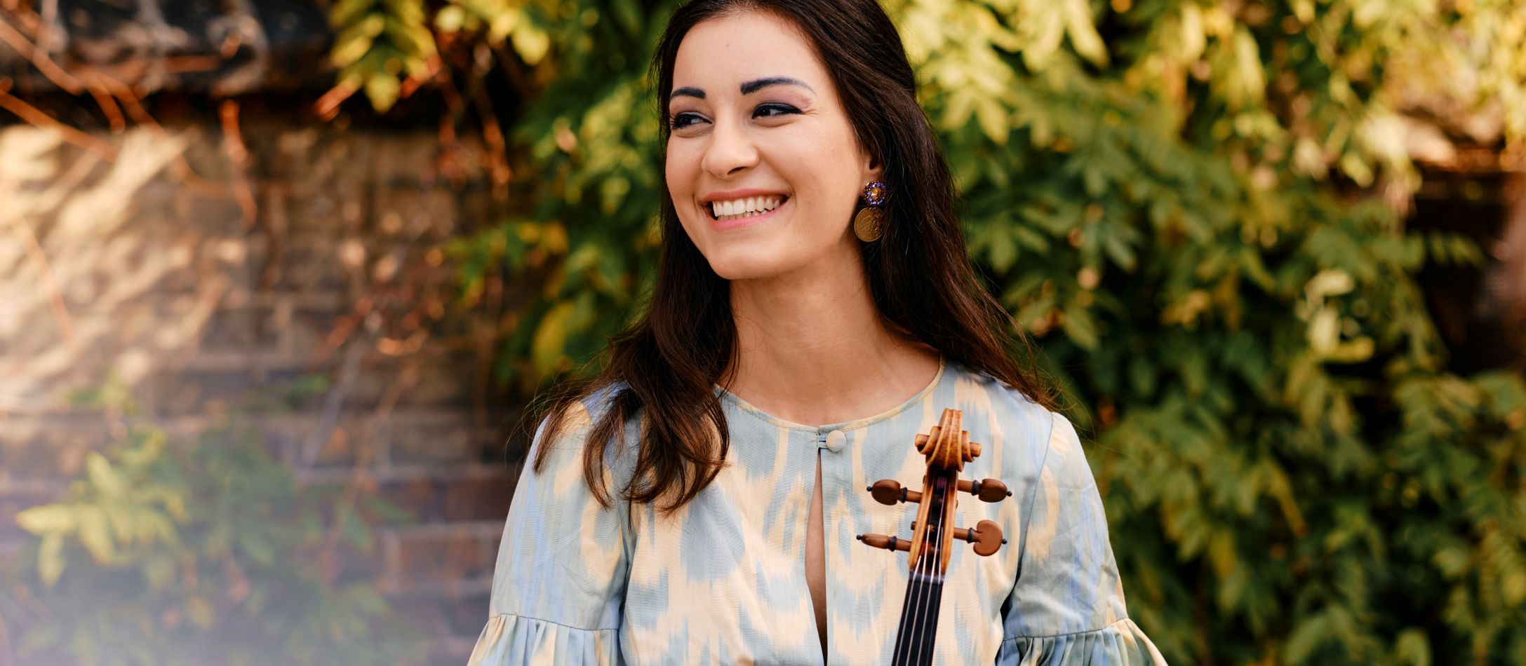 Kvinde i blå og hvid kjole, står smilende og holder sin bratsch, foran en mur og grønne grene.