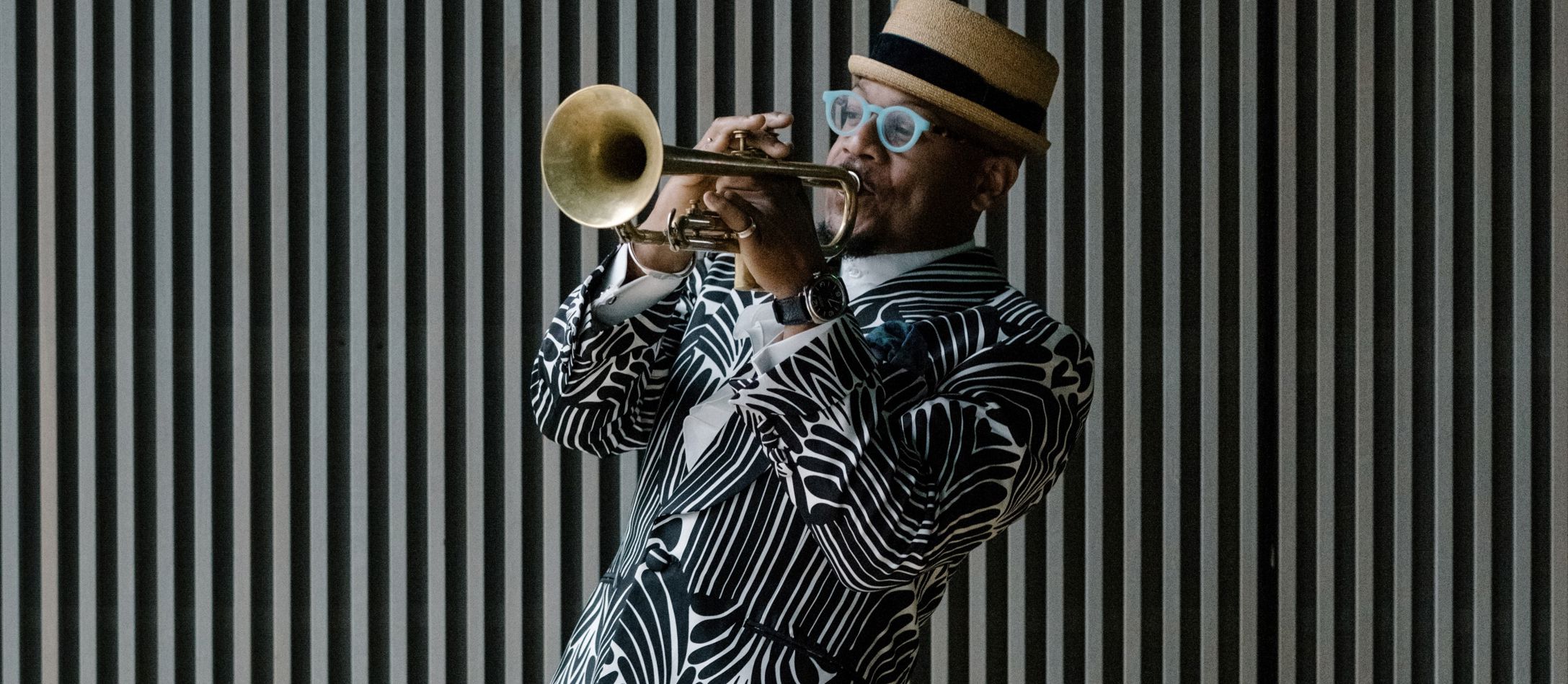 Mand i sort og hvid geometrisk blazer og hat, spiller trompet foran en sort og hvid stribet væg.