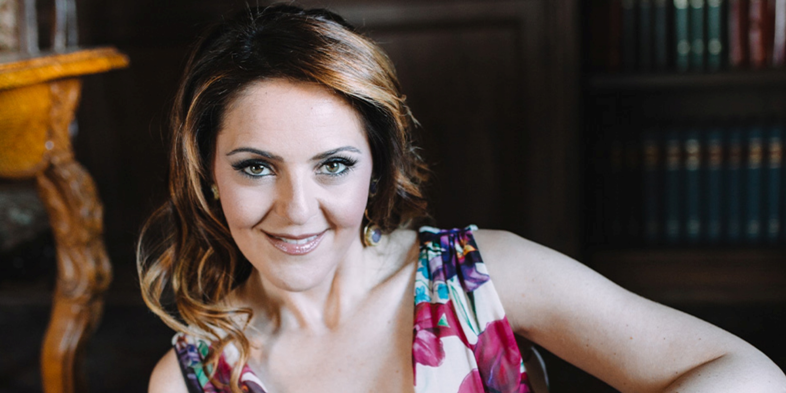 Kvinde i blomstret kjole, smiler siddende foran en bogreol.