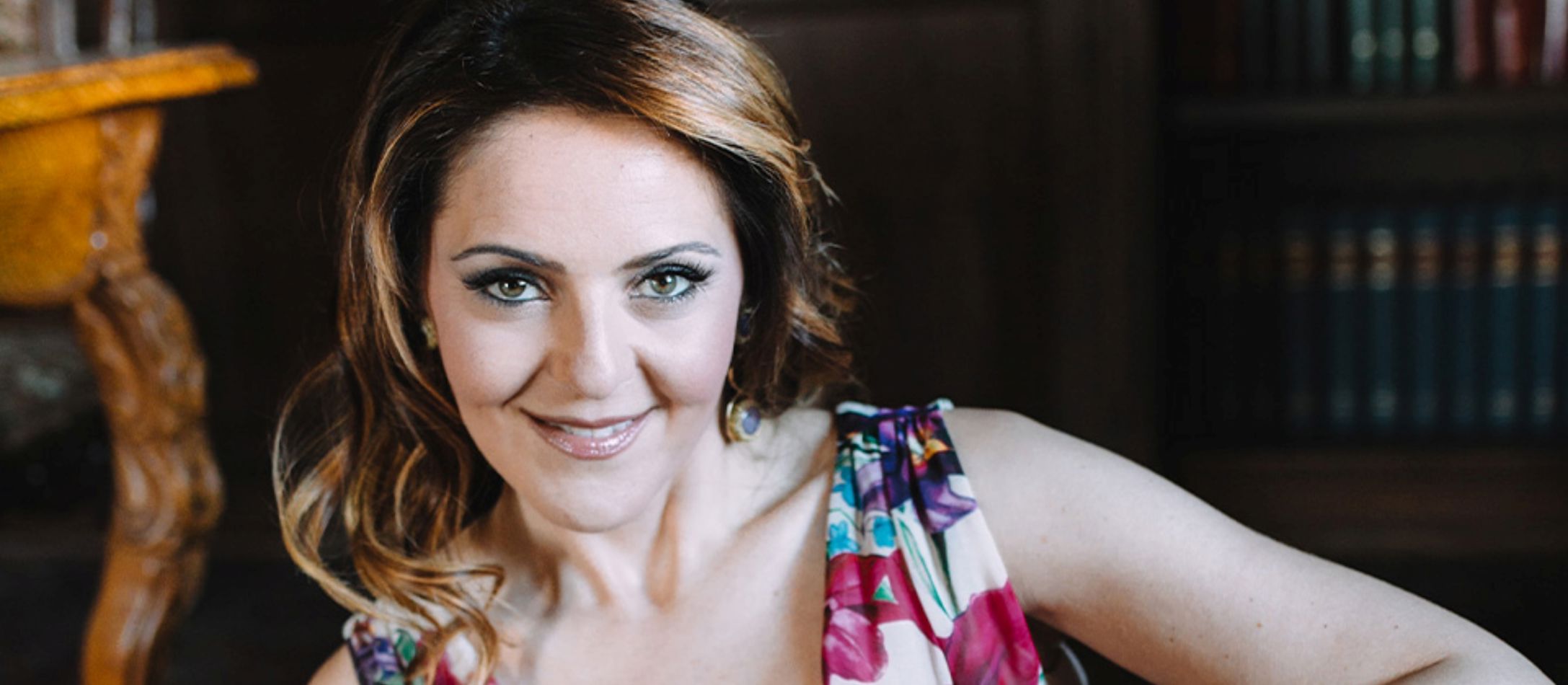 Kvinde i blomstret kjole, smiler siddende foran en bogreol.