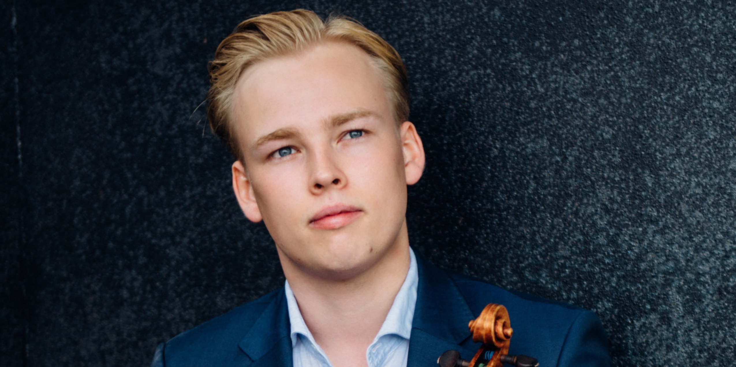 Mand i lyseblå skjorte og blå blazer med violin i hånden foran mørkeblå meleret væg