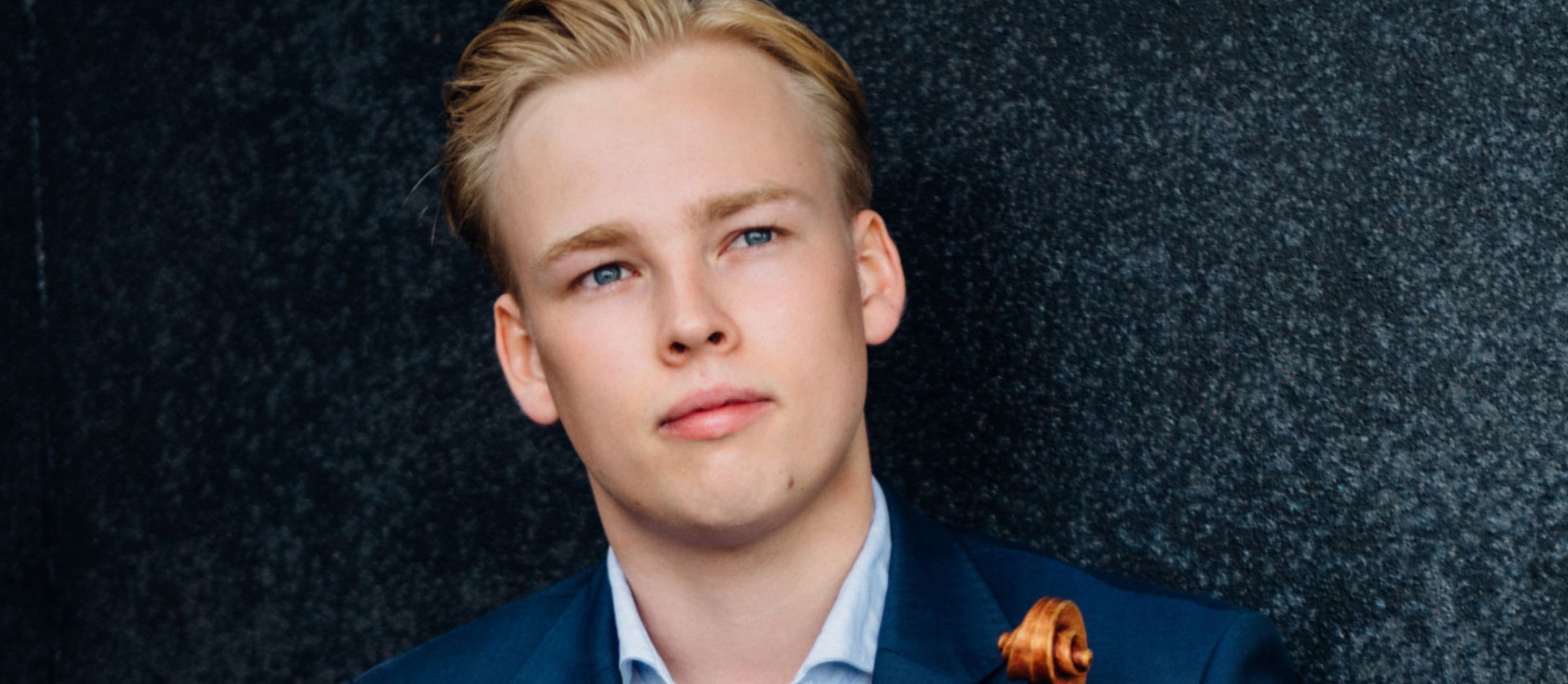 Mand i lyseblå skjorte og blå blazer med violin i hånden foran mørkeblå meleret væg