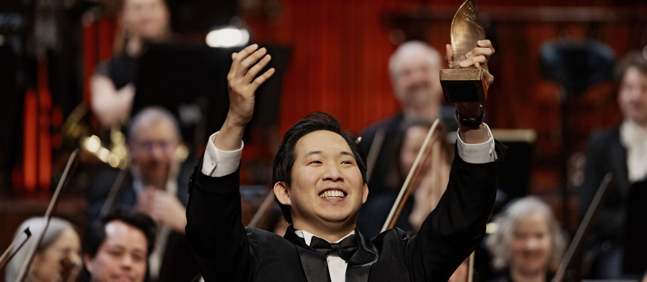 Storsmilende sydkoreanske Samuel S. Lee i klassisk suit med statuette i den ene hånd og stort orkester i baggrunden