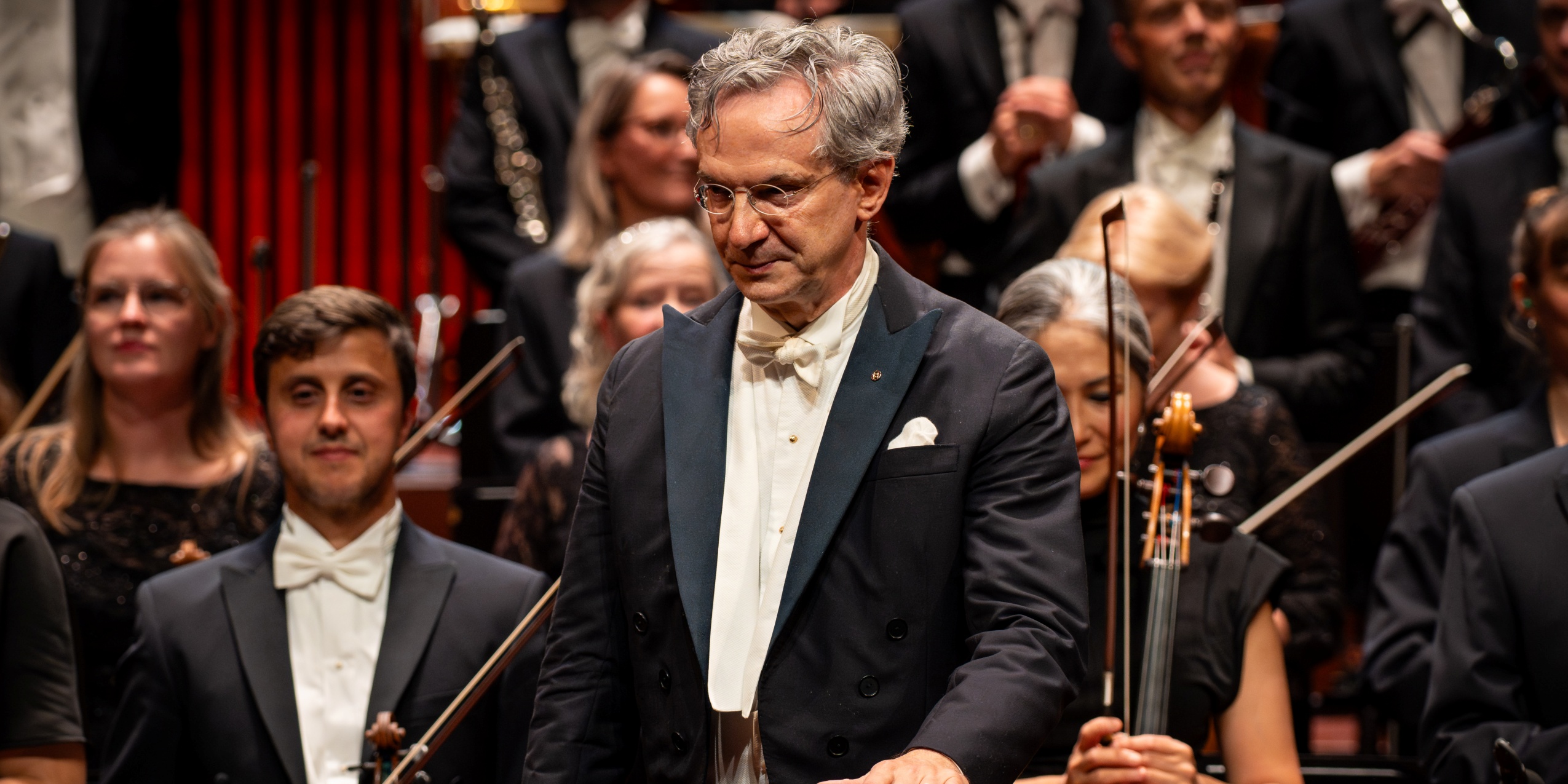 Mand i kjole og hvidt, står foran orkesteret i koncertsalen.