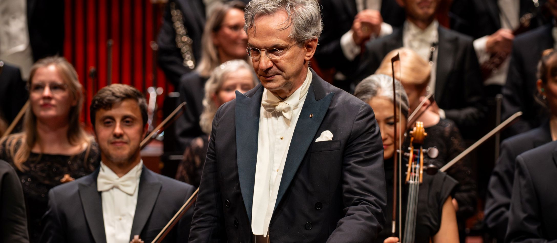 Mand i kjole og hvidt, står foran orkesteret i koncertsalen.