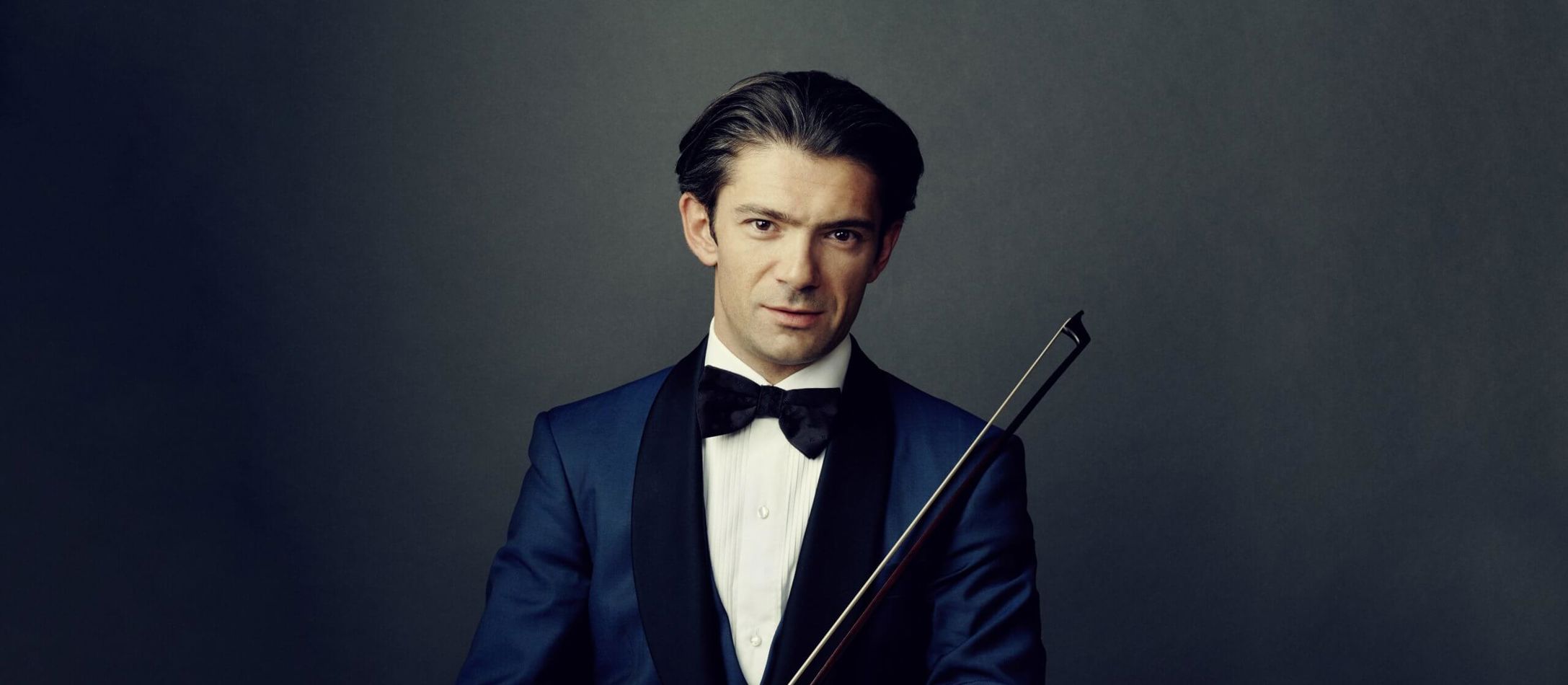 Gautier Capuçon cellostjerne iført hvid skjorte, sort butterfly og blå habitjakke foran mørkeblå baggrund
