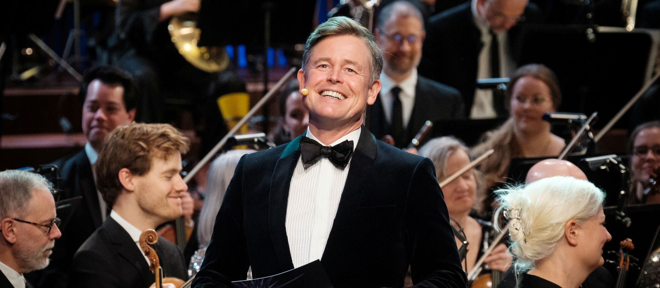 Storsmilende vært Caspar Philipson iført klassisk jakkesæt med et orkester i baggrunden.