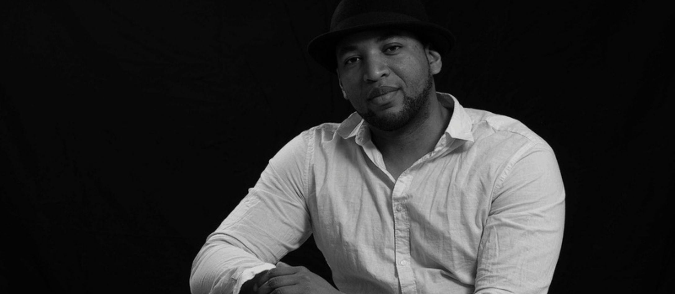 Mand i hvid skjorte og sort hat kigger ind i kameraet.