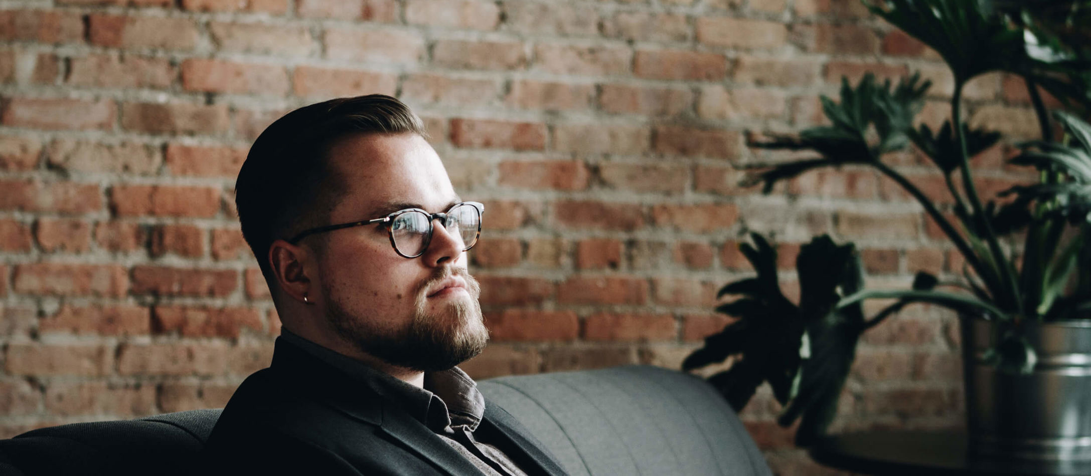 James Ley sidder i sort sofa iført briller med flot skæg foran rød murstensvæg indendørs