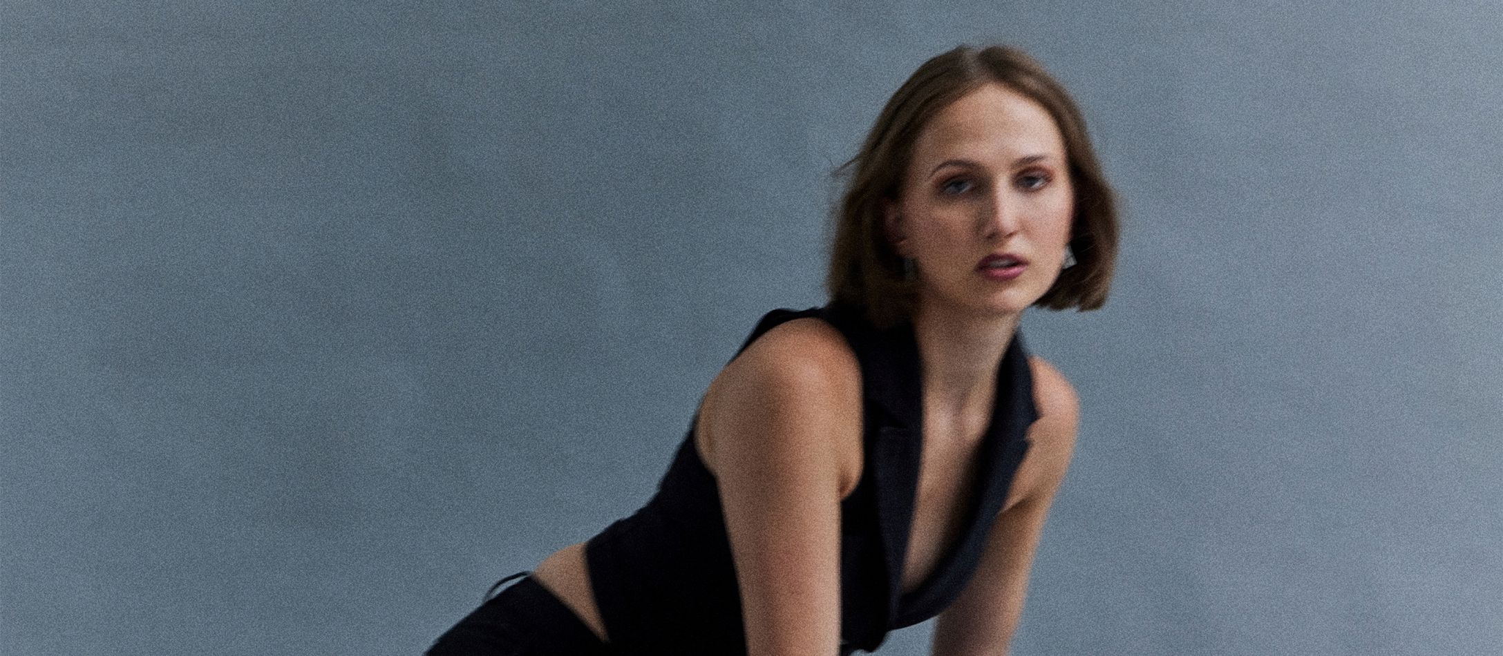 Sløret CEVIL med mørkt pagehår og intense mørke øjne iført habitskjorte uden armer på grålig baggrund.