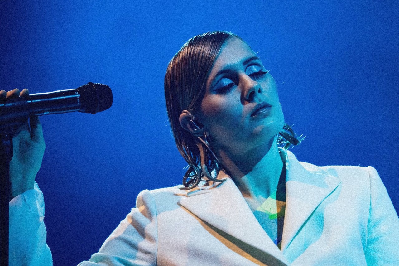 Eee Gee med lukkede øjne i blåt skært og blå baggrund iført hvidt jakkesæt og slikket hår og fuld makeup. 