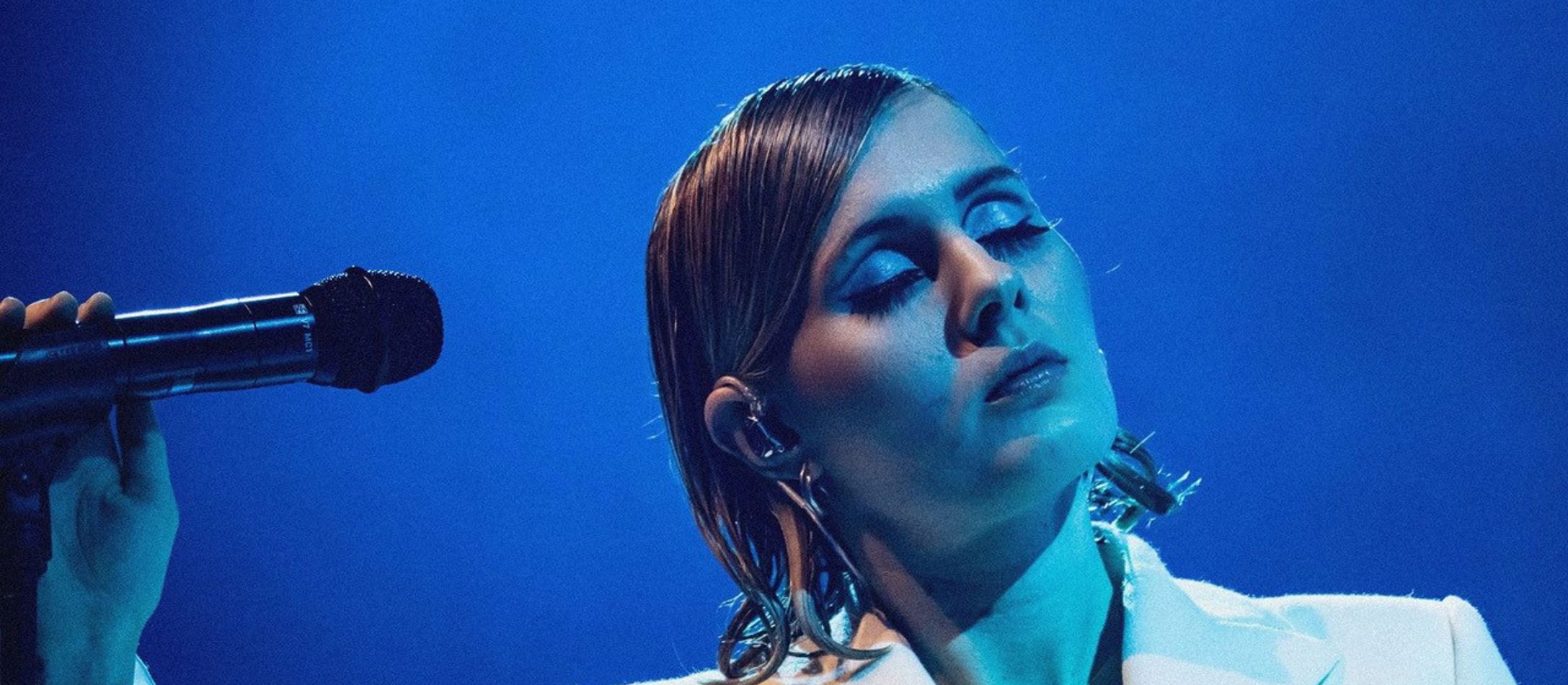 Eee Gee med lukkede øjne i blåt skært og blå baggrund iført hvidt jakkesæt og slikket hår og fuld makeup.