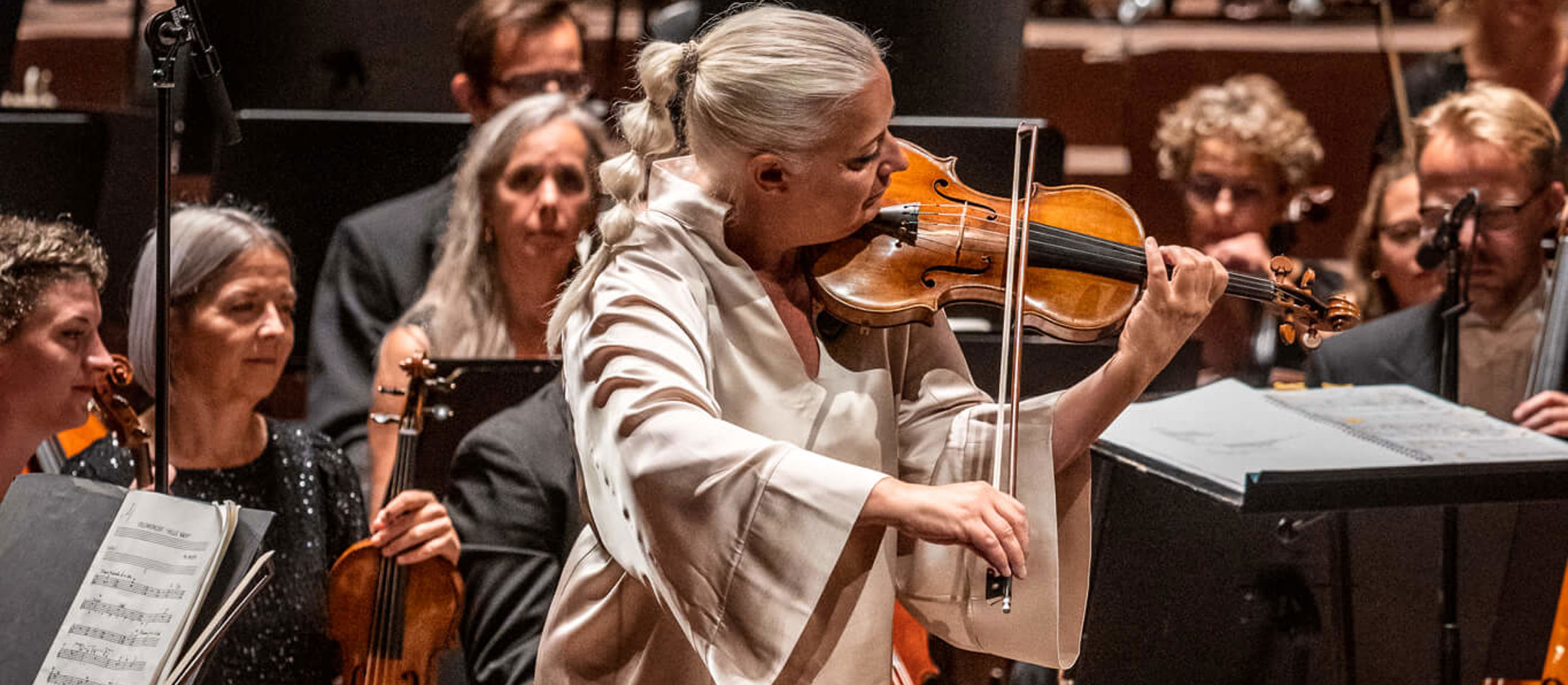 Christina Åstrand står og spiller violin i hvidt sæt tøj foran stort orkester, der sidder ned og venter.