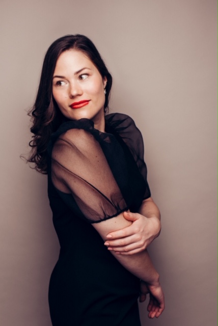 Sanger Clara Thomsen med småt krøllet mørkt hånd og røde læber iført sort trøje står med siden til foran lys baggrund. 