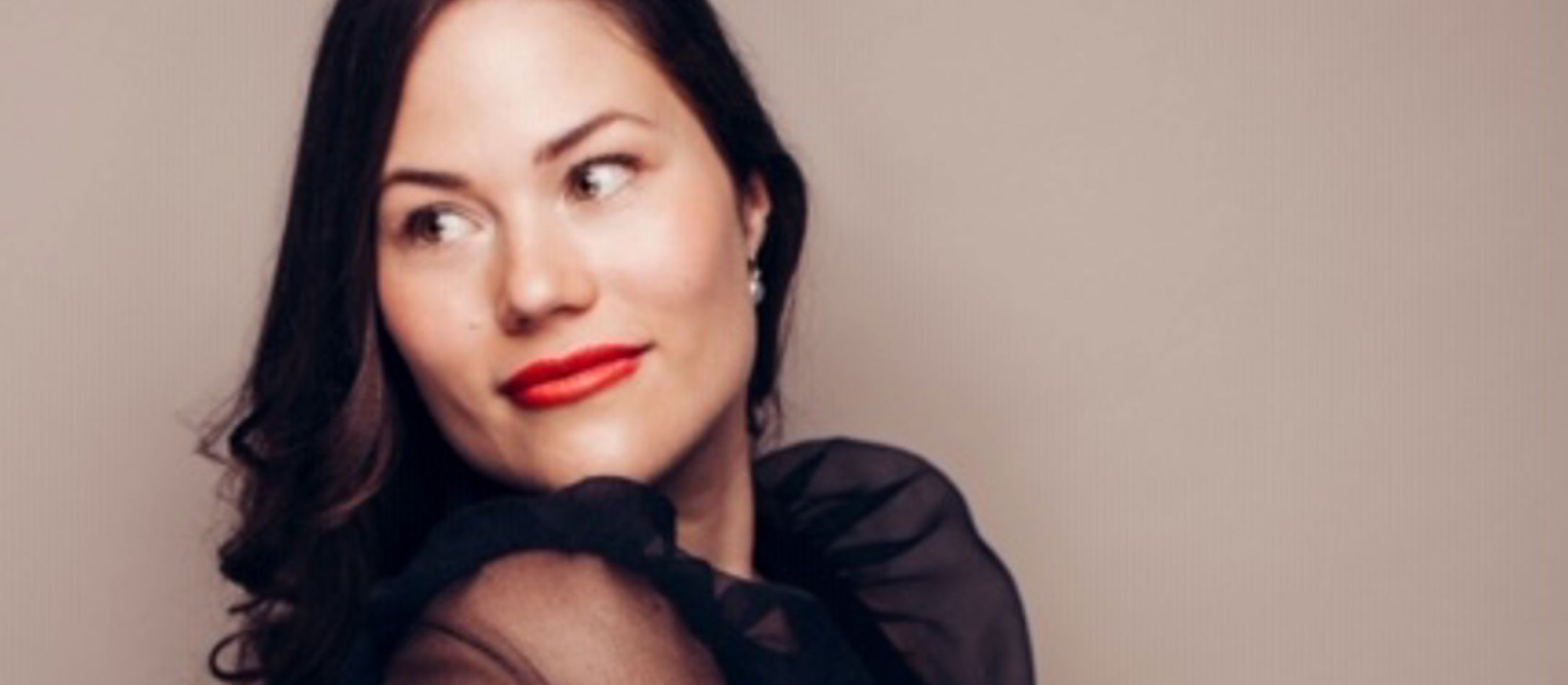 Sanger Clara Thomsen med småt krøllet mørkt hånd og røde læber iført sort trøje står med siden til foran lys baggrund.