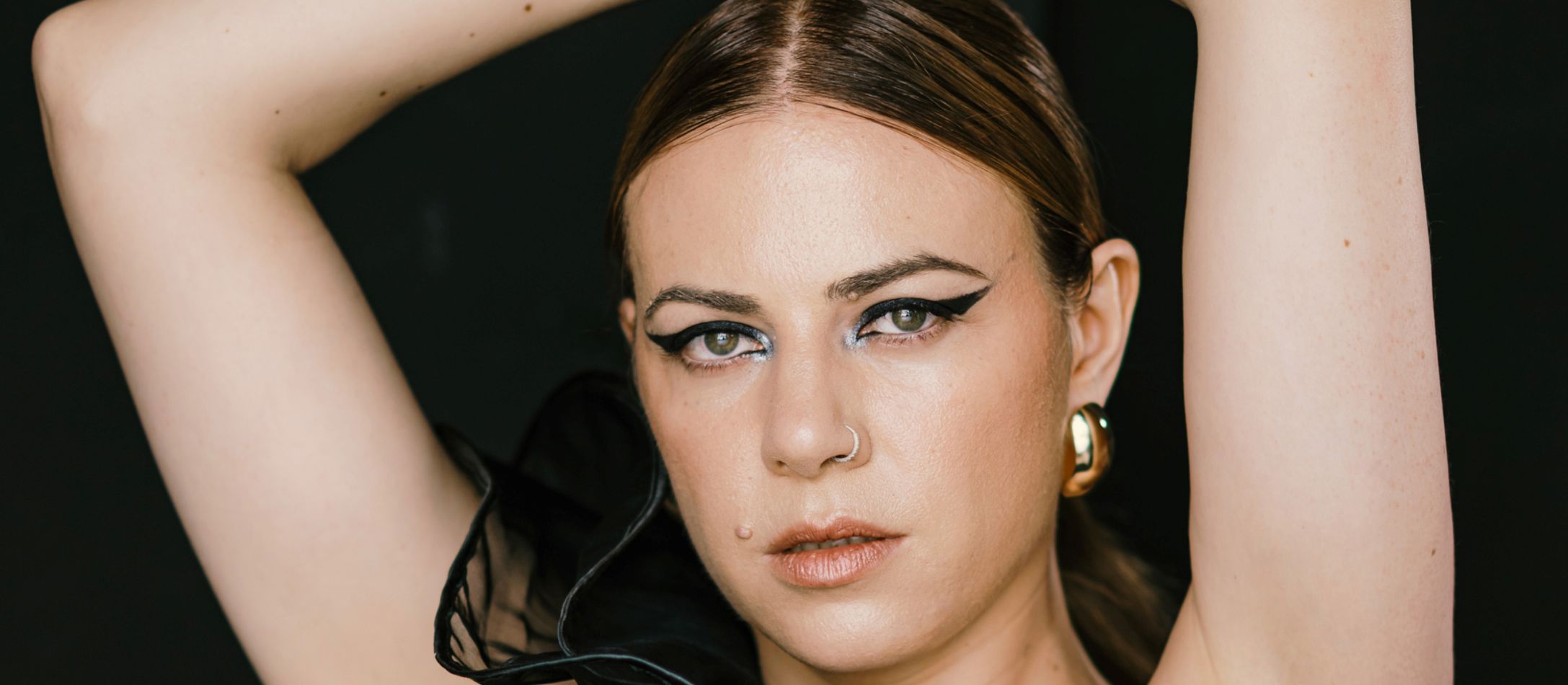 Kvinde i sort kjole med hænderne oppe over hovedet foran sort baggrund