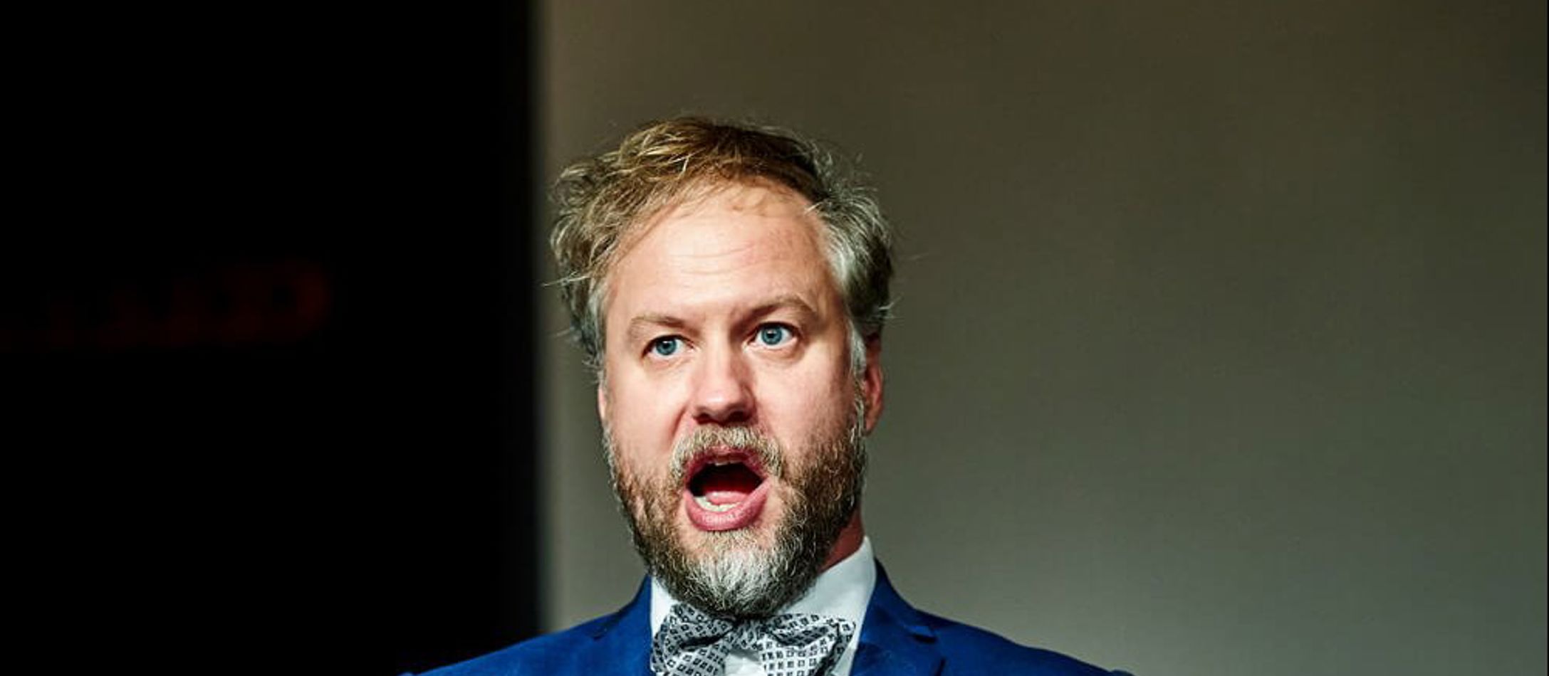 Andreas Landin synger med åben mund og holder en sangbog ud i hænderne foran sig iført hvid skjorte, butterfly og blå jakke.