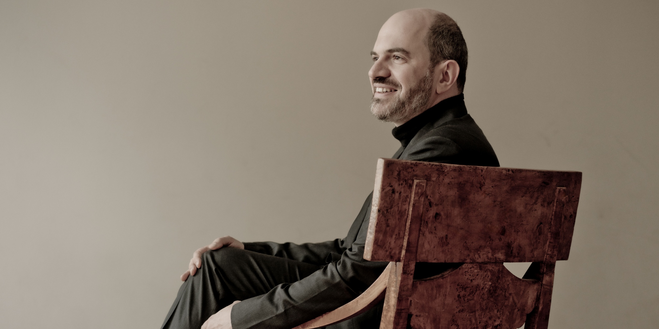 Man i sort, sidder smilende i en stor træstol foran beige væg.