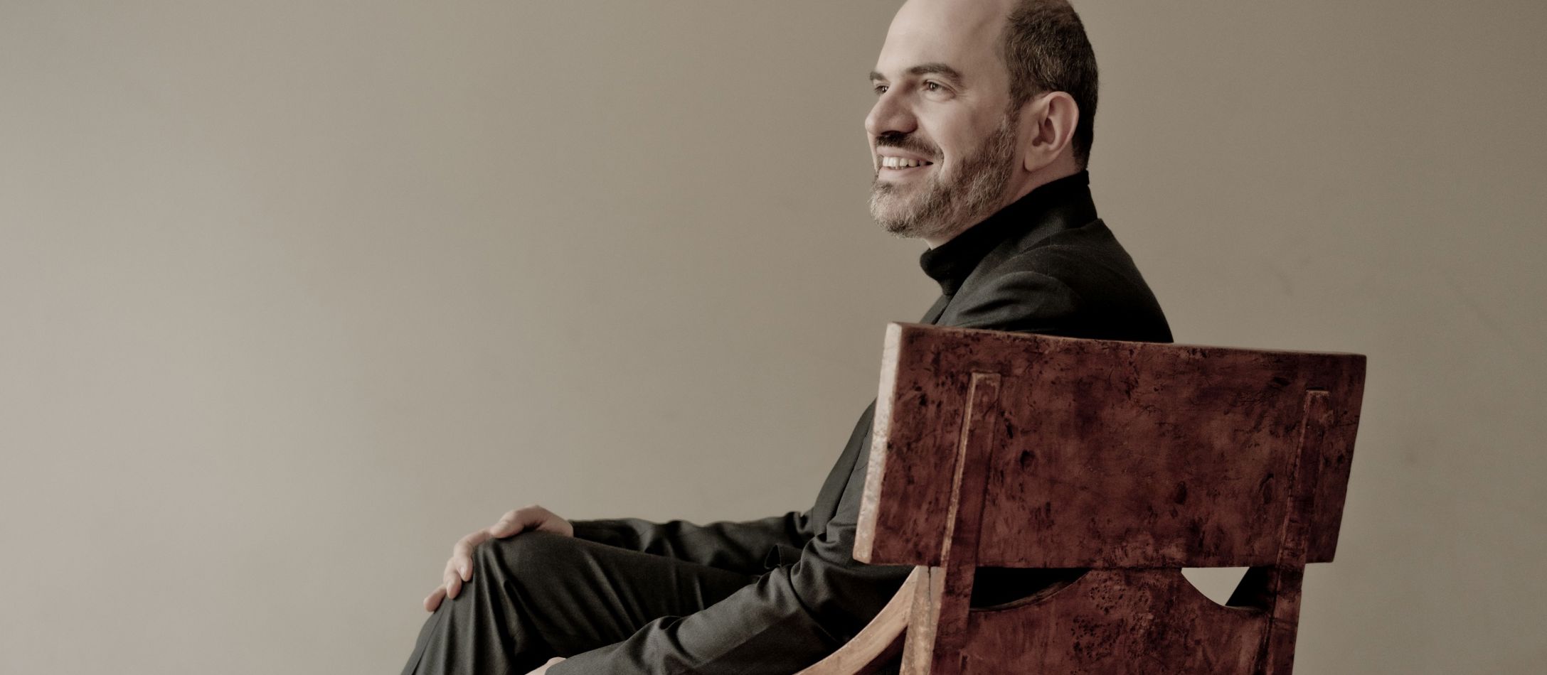 Man i sort, sidder smilende i en stor træstol foran beige væg.