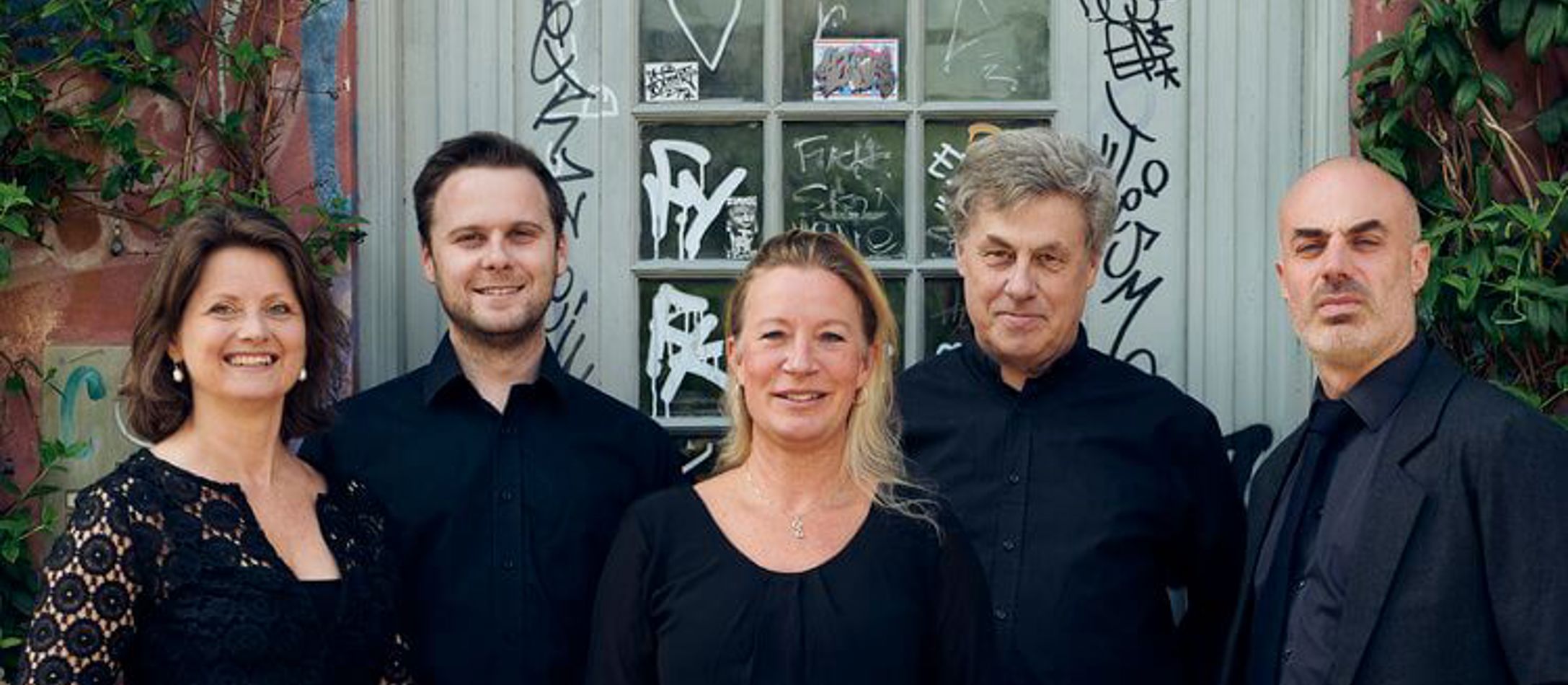 Gruppebillede af 2 kvinde og tre mænd fra Theatre Of Voices iført pænt sort tøj foran grå døren og grønne buske på brosten.