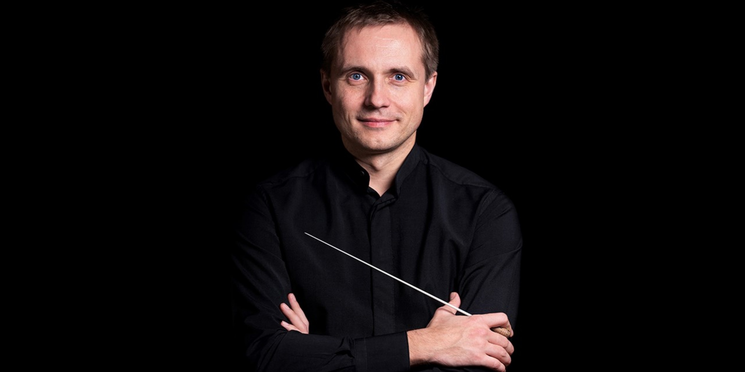 Mand med dirigentstok og sort skjorte kigger ind i kameraet foran sort baggrund