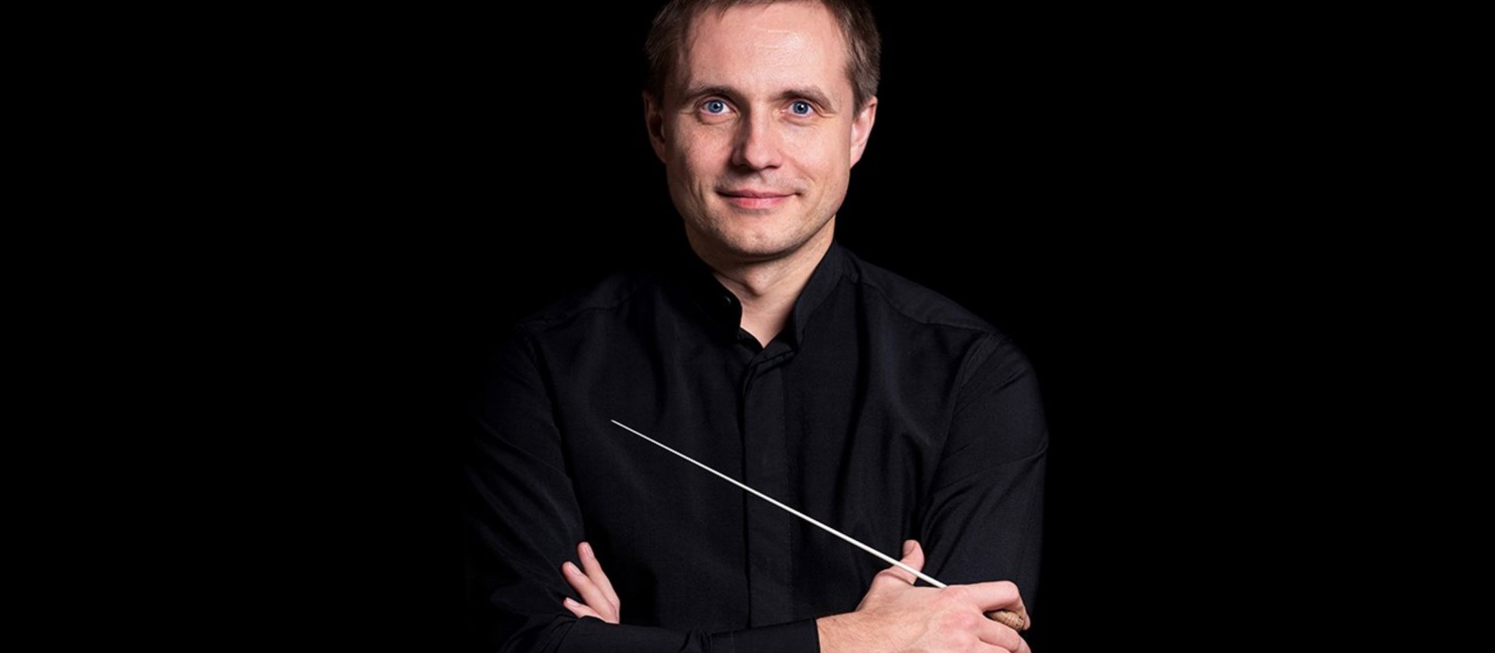 Mand med dirigentstok og sort skjorte kigger ind i kameraet foran sort baggrund