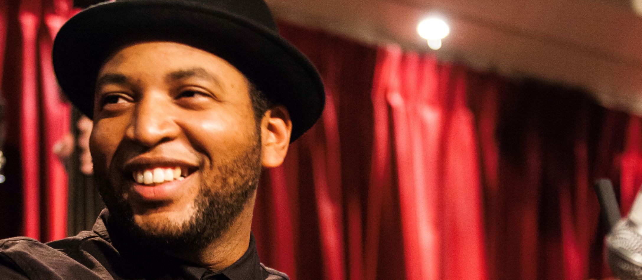 Eliel Lazo iført sort bowlerhat og sortknappet skjorte smiler varmt foran røde satingardiner.