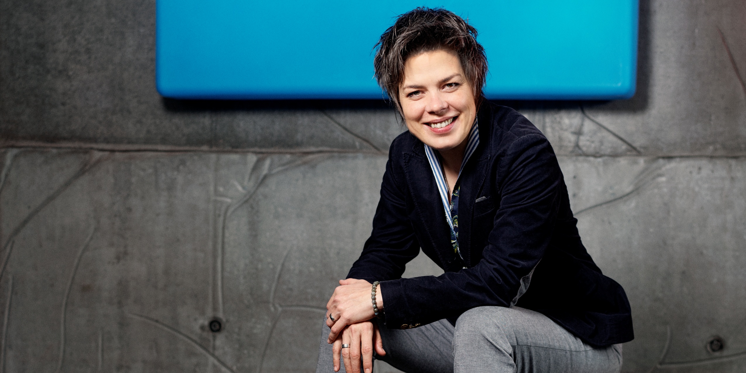 Kvinde kigger smilende ind i kameraet, siddende foran grå betonvæg med en blå væglampe.