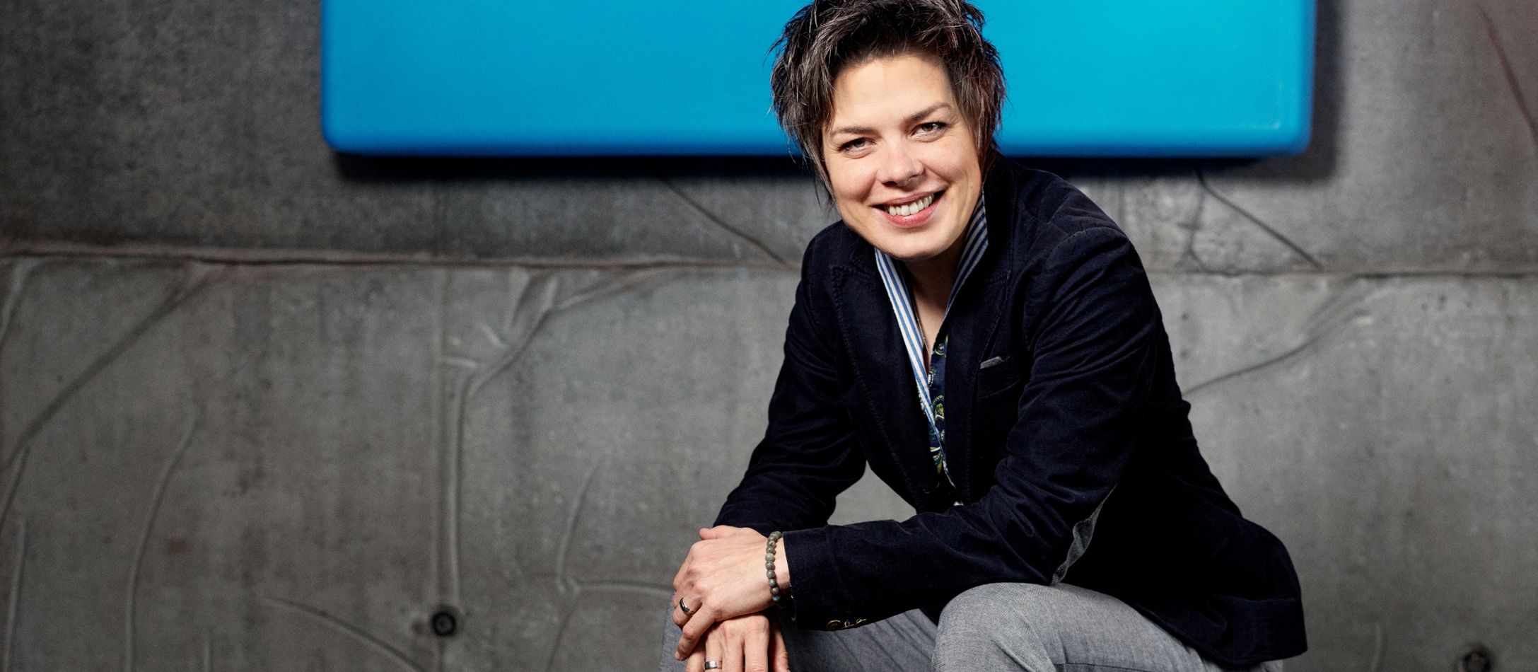Kvinde kigger smilende ind i kameraet, siddende foran grå betonvæg med en blå væglampe.