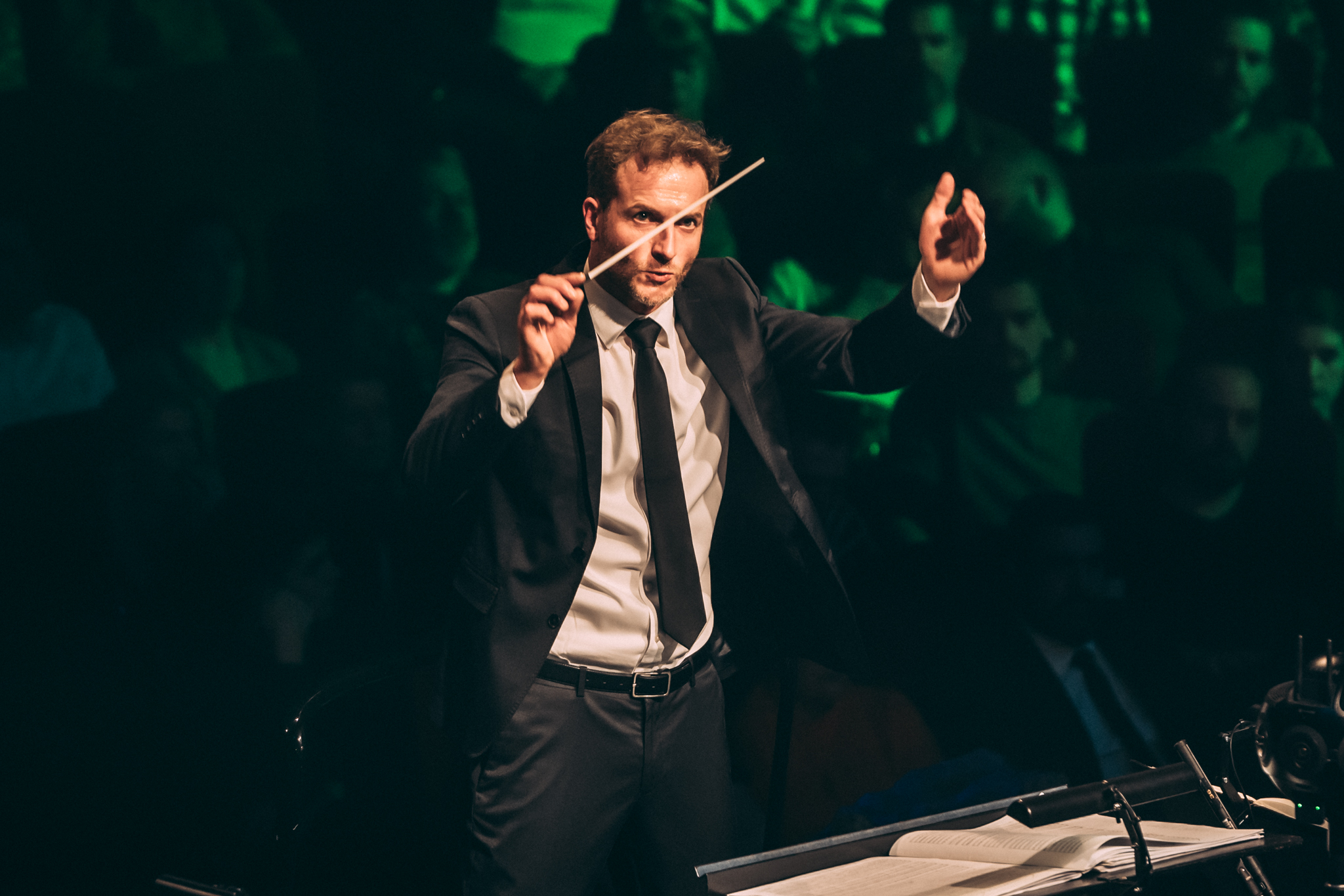 Dirigent Christian Schuman iført klassisk jakkesæt løfter dirigentstang i færd med at dirigere et orkester.