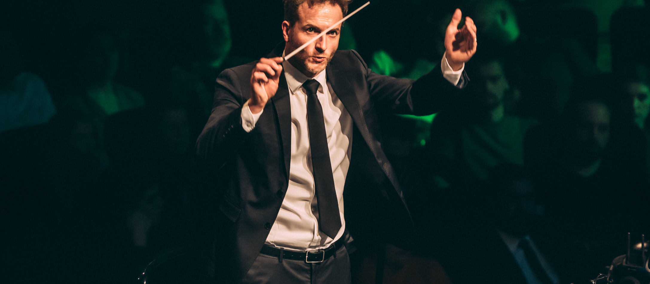 Dirigent Christian Schuman iført klassisk jakkesæt løfter dirigentstang i færd med at dirigere et orkester.