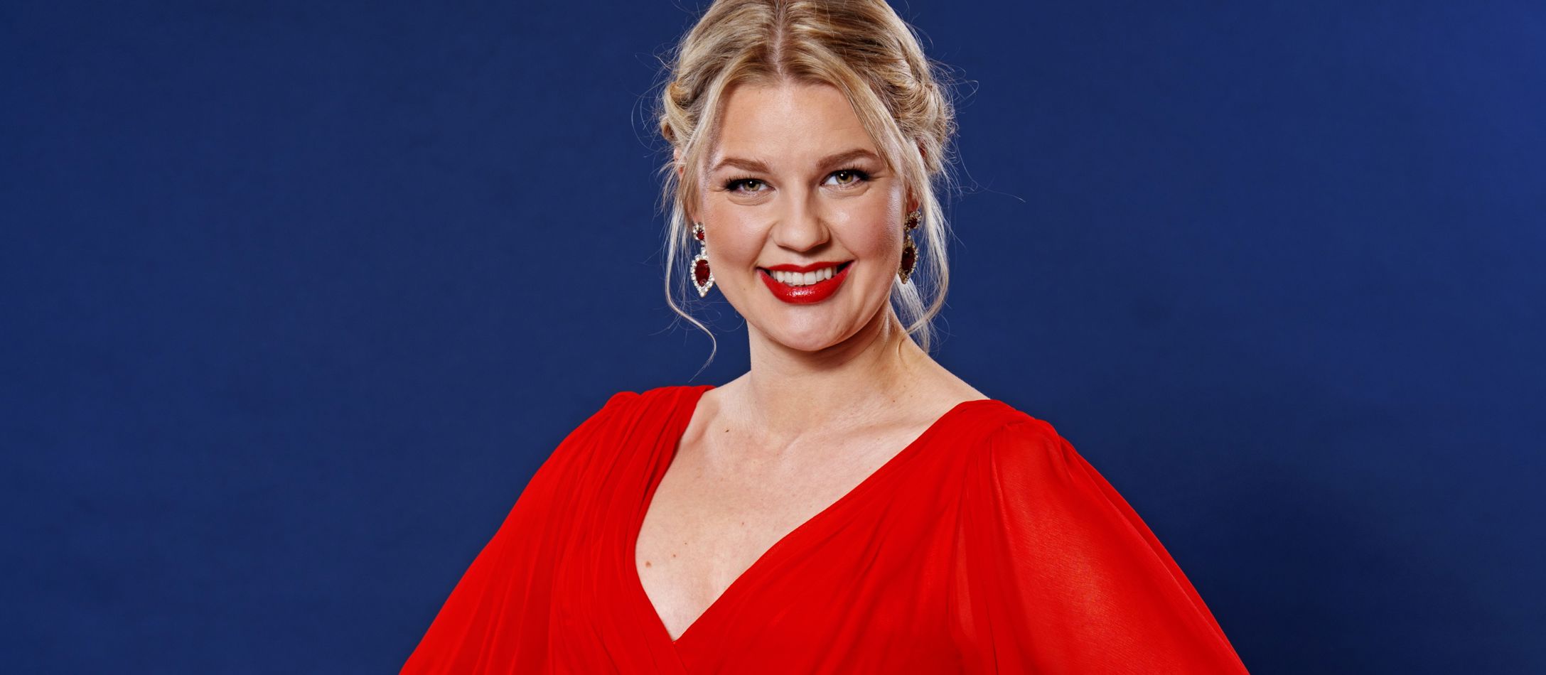 Kvinde i rød kjole, kigger ind i kameraet foran blå baggrund.