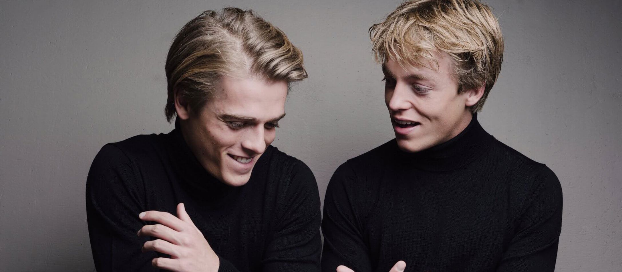 De lyshårede klaverbrødre Lucas & Arthur Jussen begge iført sort halskrave sidder ved reflekterende klaver og smiler til hinanden.