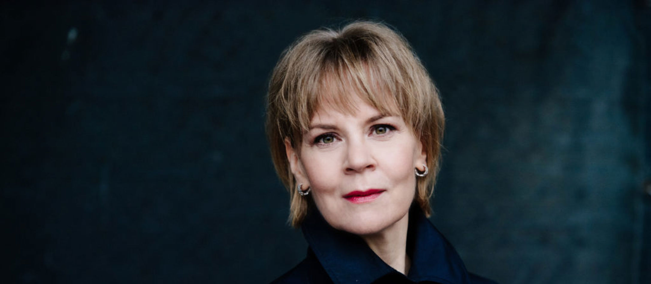 Dirigenten Susanna Mälkki med skulderkort lyst hår iført lukket mørkeblå efterårsjakke foran mørk baggrund.